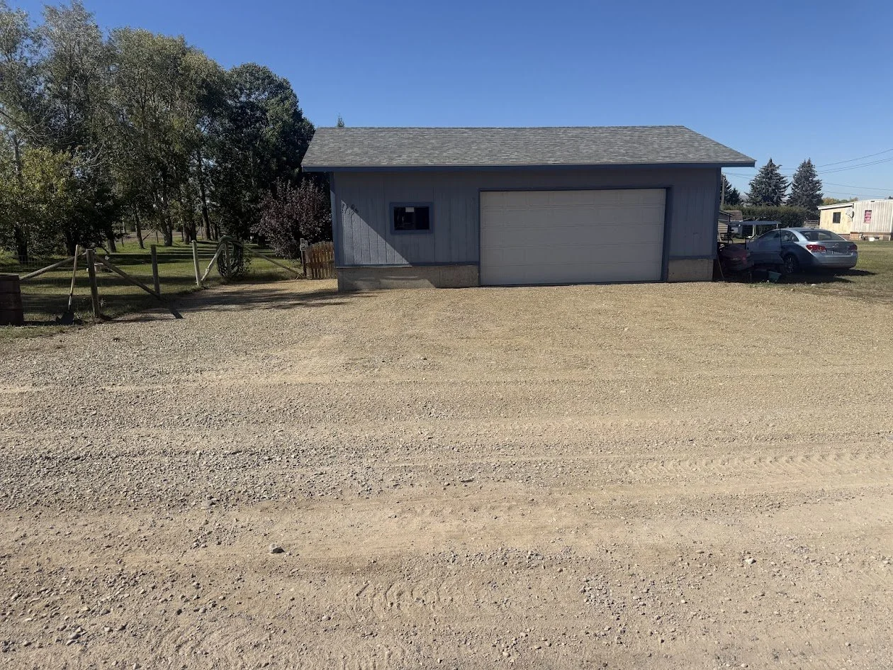 A small blue building with a gray garage door and a small window, situated on a gravel lot with some trees and cars in the background.
