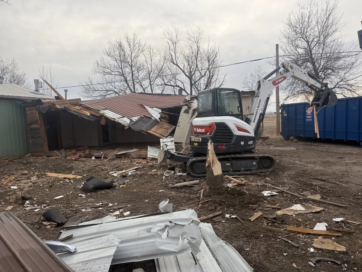 A construction mini excavator demolishing a small wooden building with debris scattered on the ground, including metal roofing sheets and broken wood pieces.
