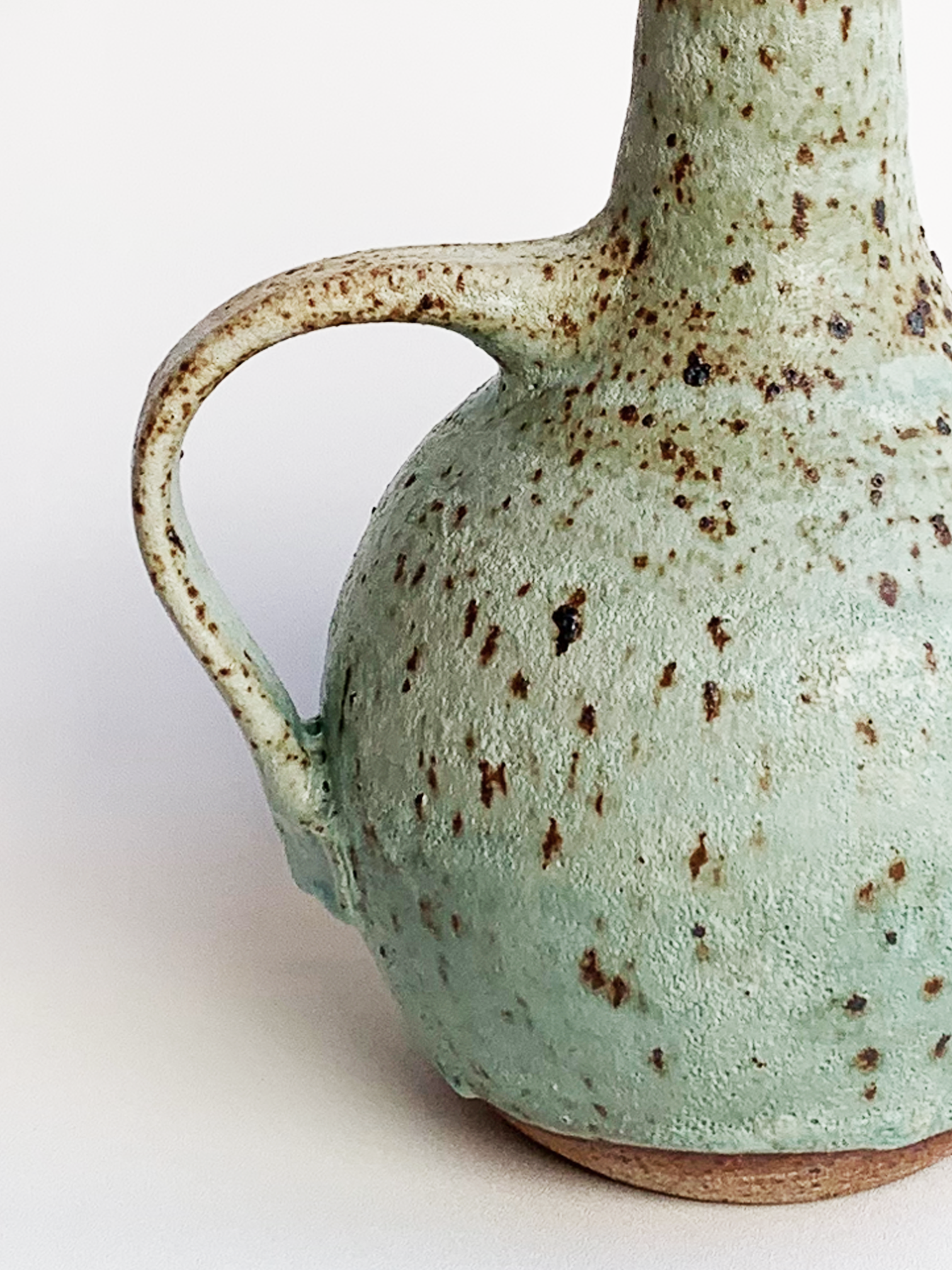 Handmade stoneware oil cruet with a turquoise speckled glaze, rounded body, curved handle, and metal weighted pouring spout.