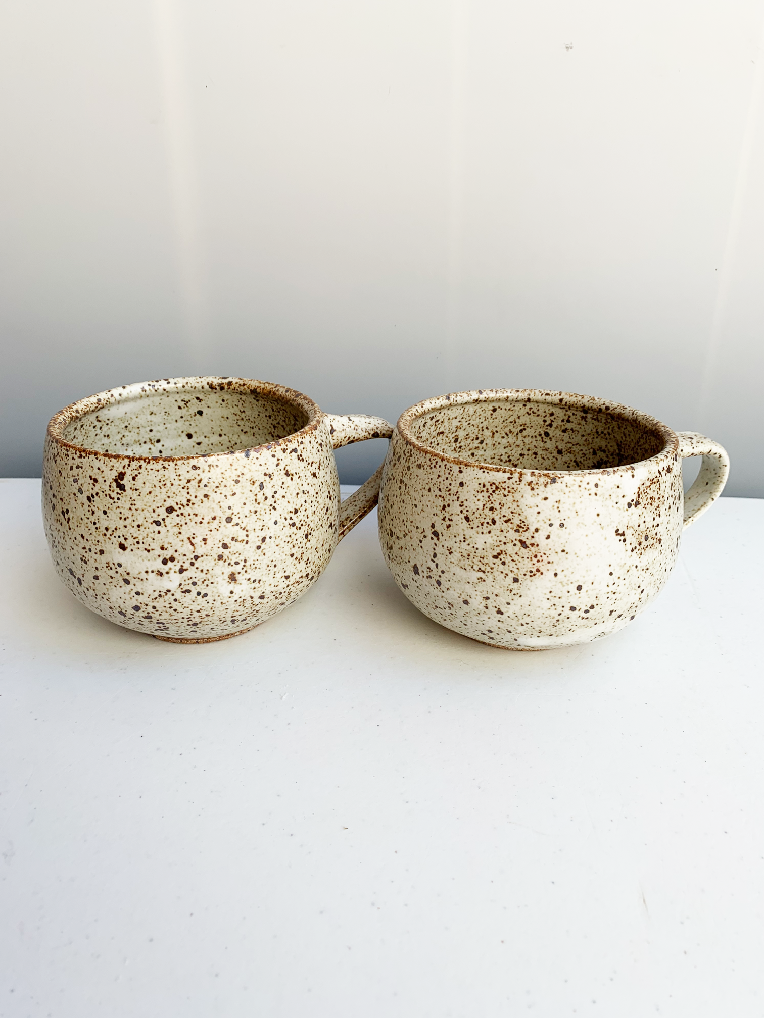 Handmade round stoneware mug with a pale speckled glaze, brown flecks, curved body, and side handle.