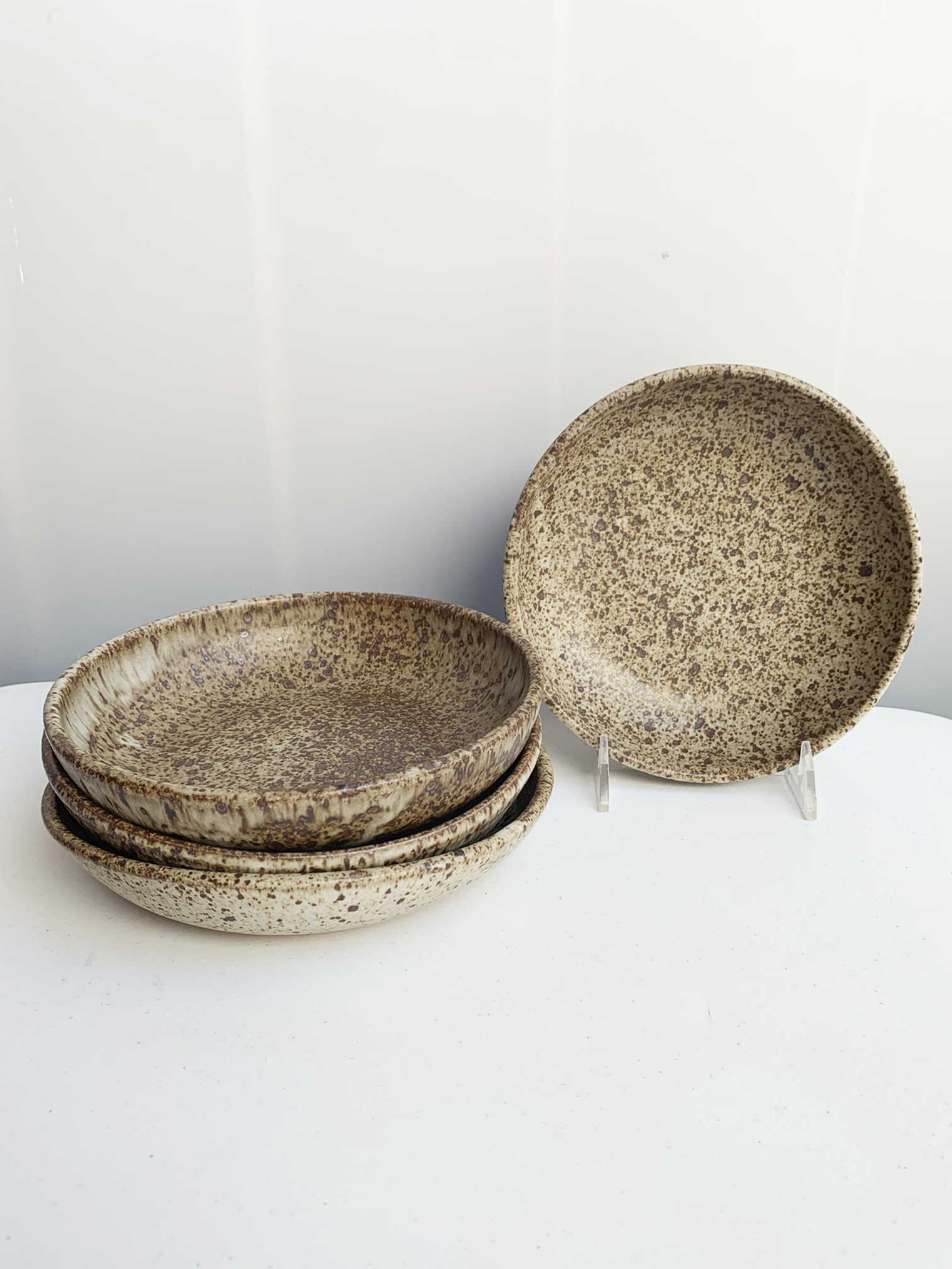 Stack of handmade stoneware pasta bowls with a warm speckled glaze and gently raised sides, with one bowl displayed upright.