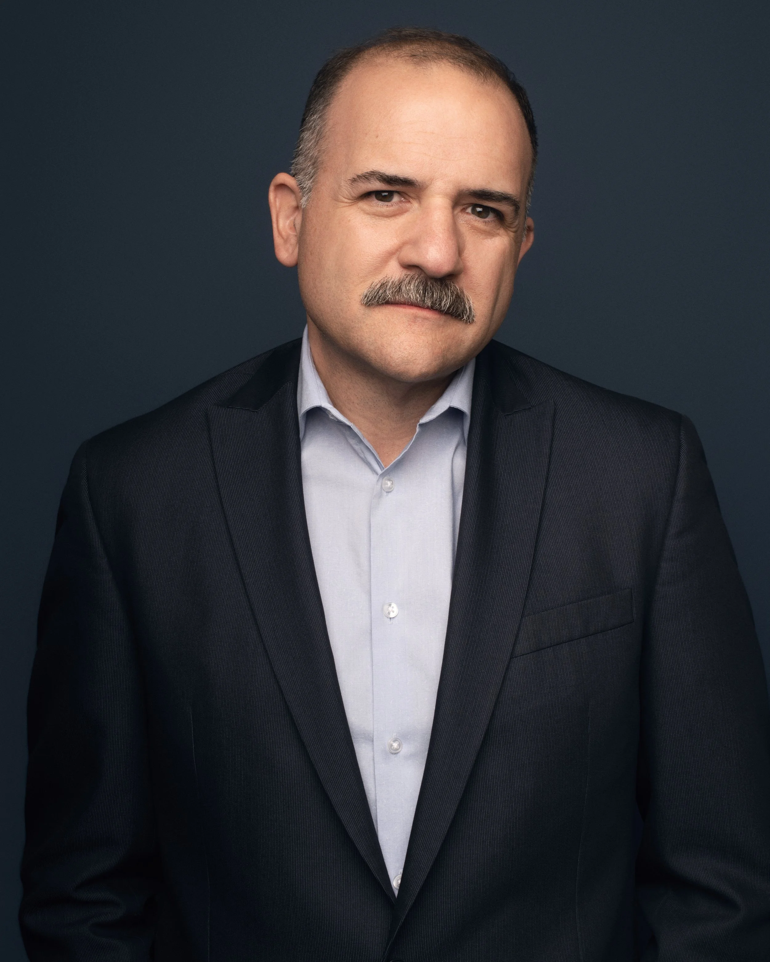 A middle-aged man with a mustache and short hair, wearing a dark suit jacket and a light-colored shirt, posing against a dark background.