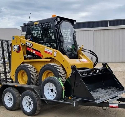 A yellow Caterpillar skid-steer loader on a black trailer with white wheels, parked outside a large industrial building.