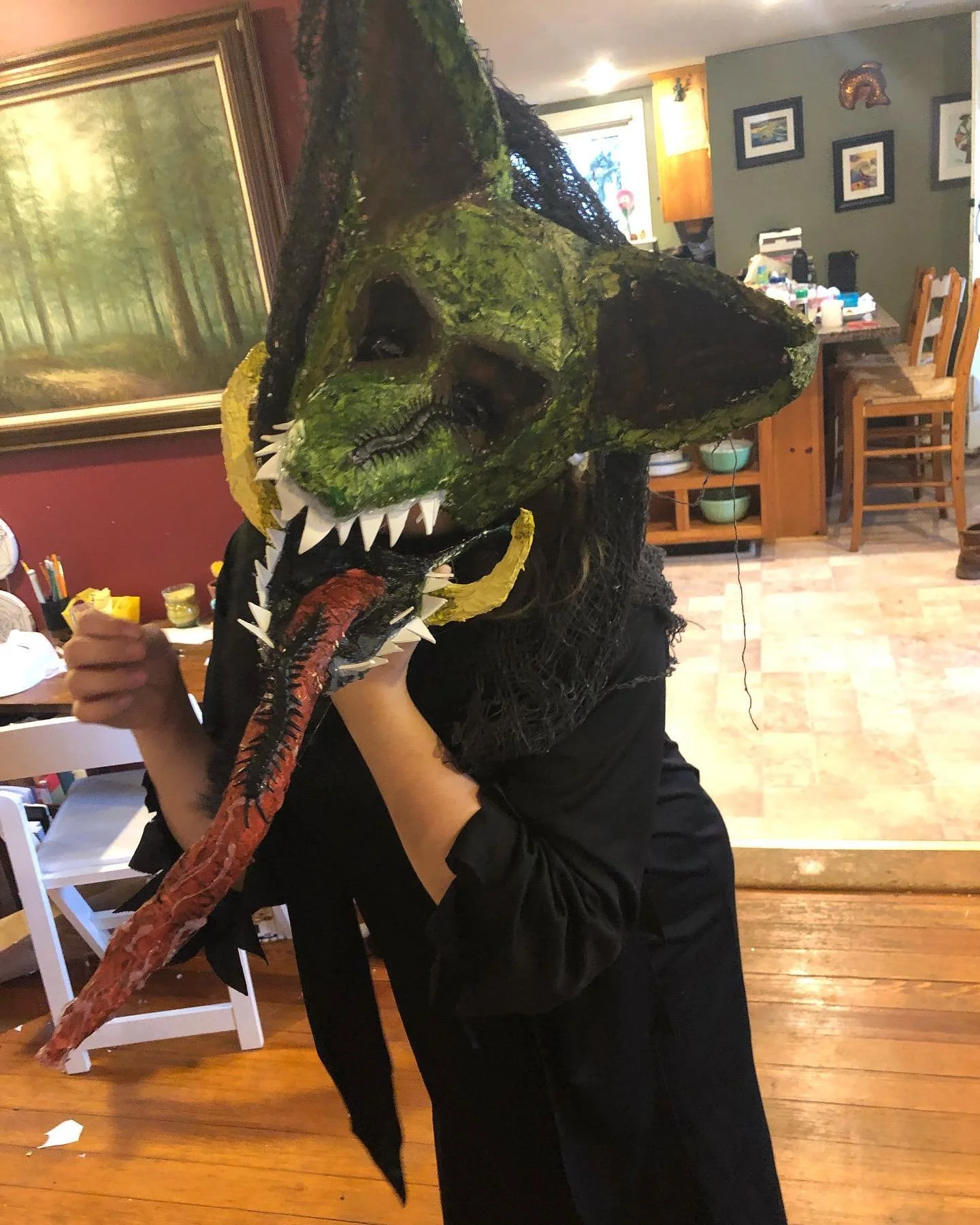 Person holding a large dinosaur skull mask with green moss-like texture, sharp teeth, and long red tongue in a room with wooden furniture and artwork on the wall.