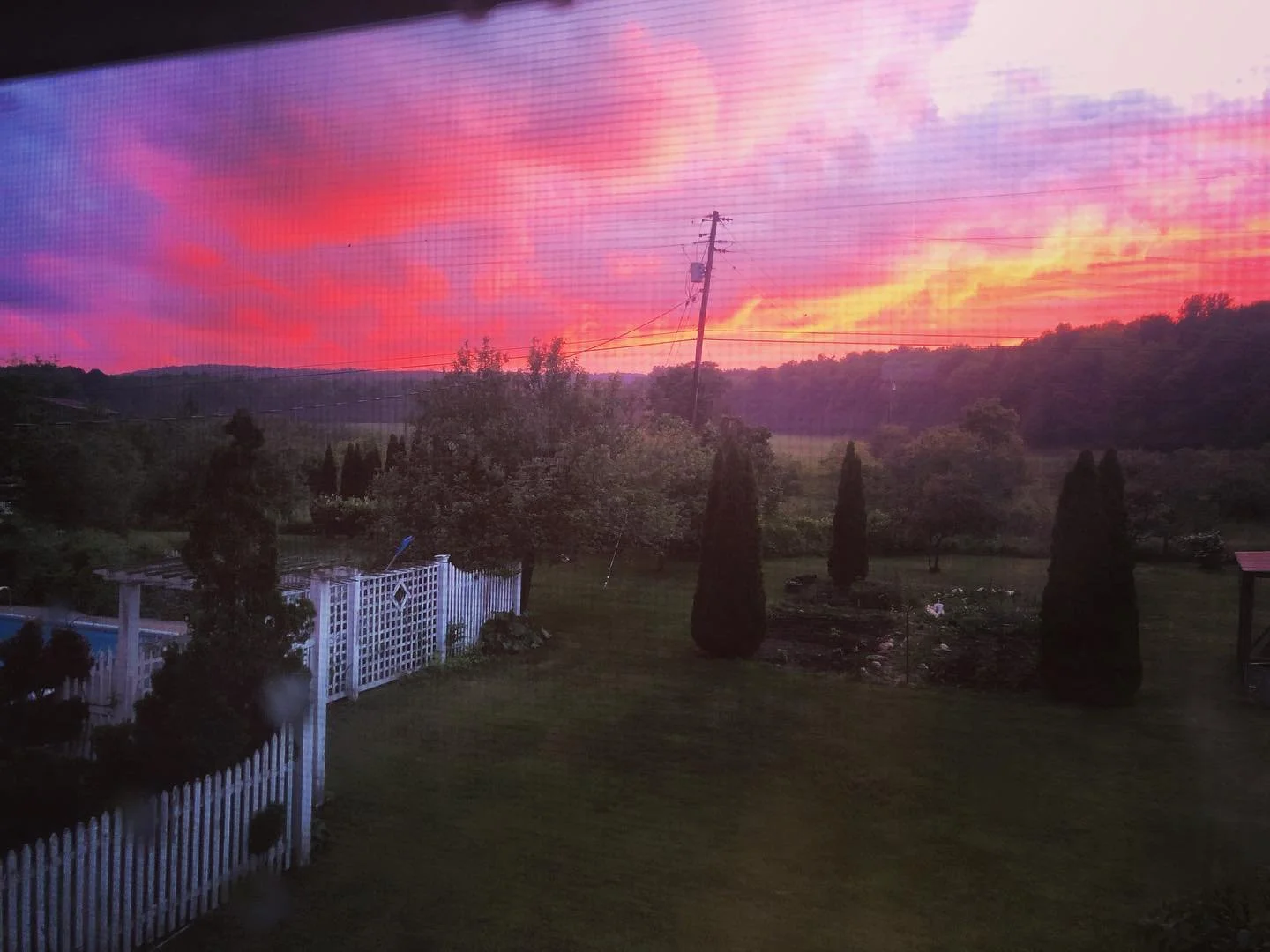 View of a sunset with vibrant pink and purple clouds over a countryside landscape, including trees, a white fence, and rolling hills.
