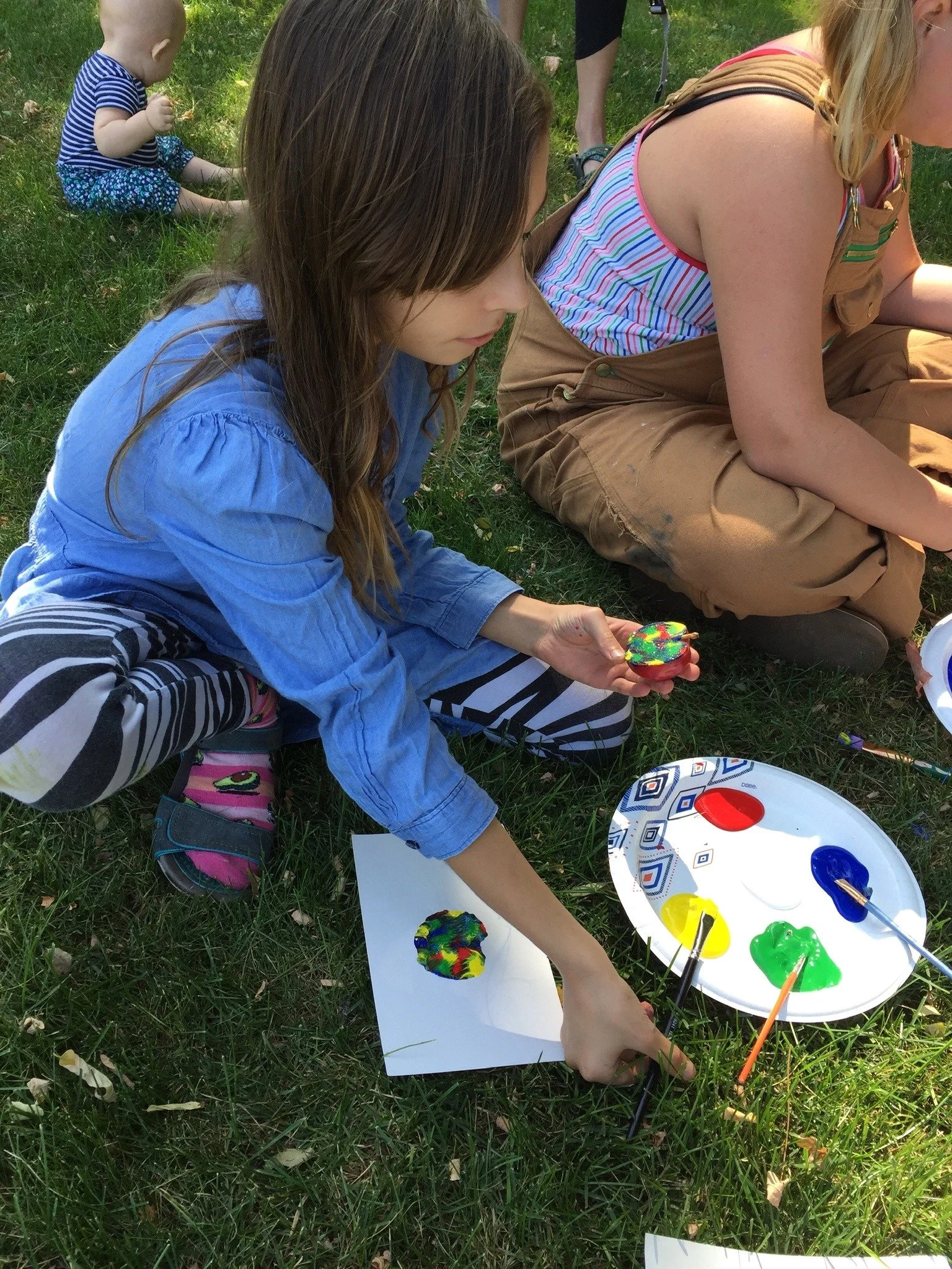 Girl with long brown hair painting a rock with colorful paint outdoors on the grass, with a plate of various paint colors nearby.