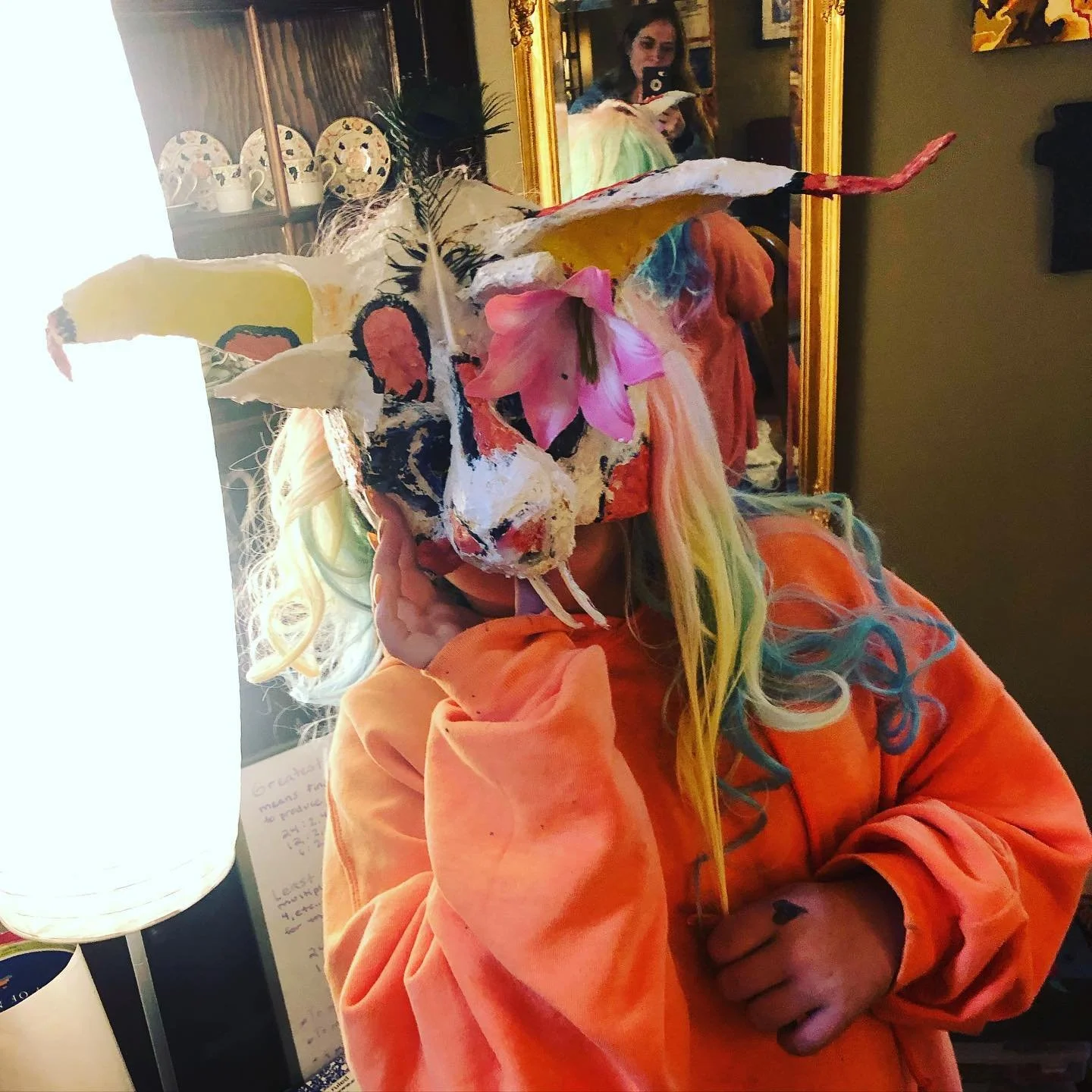Person in an orange hoodie wearing a colorful mask with feathers, flowers, and painted details, standing indoors in front of a mirror, with colorful hair and holding their hand to their face.