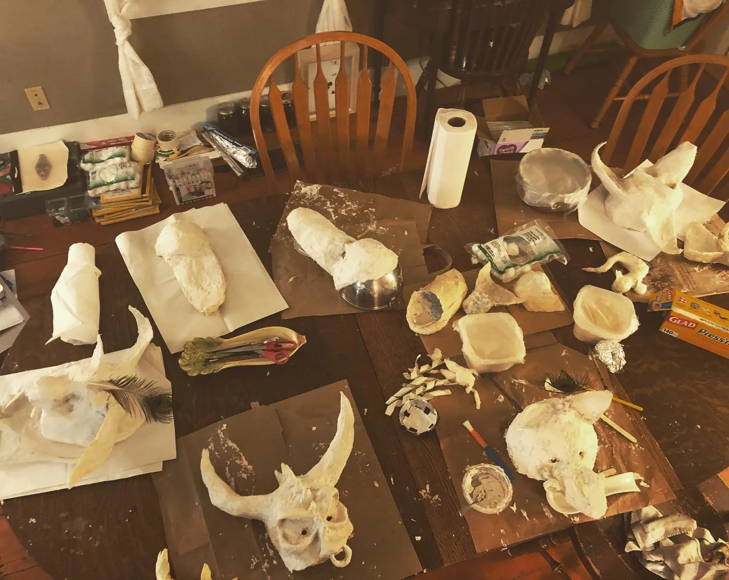 A wooden dining table covered with animal skulls, bones, and craft supplies, including paper, scissors, tape, and paint, suggesting a skull crafting or painting project.