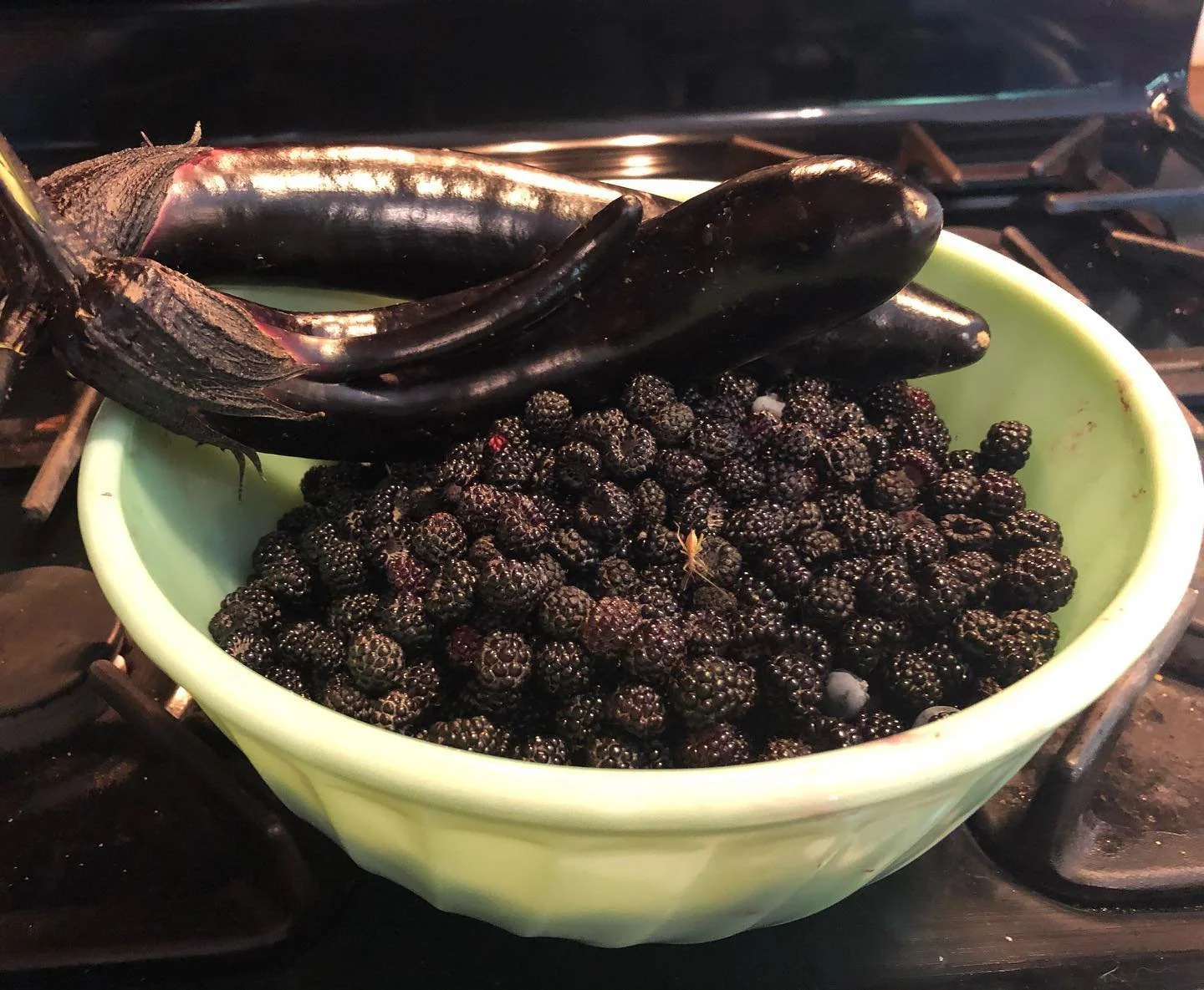 A green bowl filled with blackberries and a few eggplants on top, sitting on a stove with a burner grate visible.