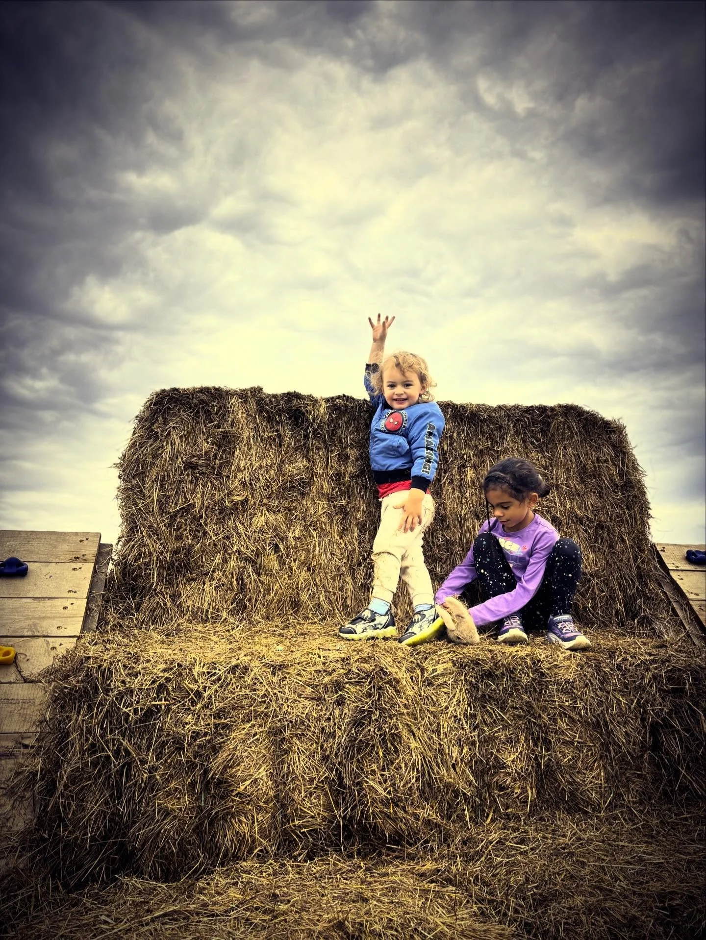 Two young children playing on a large haystack outdoors under a cloudy sky, one child standing with one arm raised and smiling, the other sitting and playing with a toy.