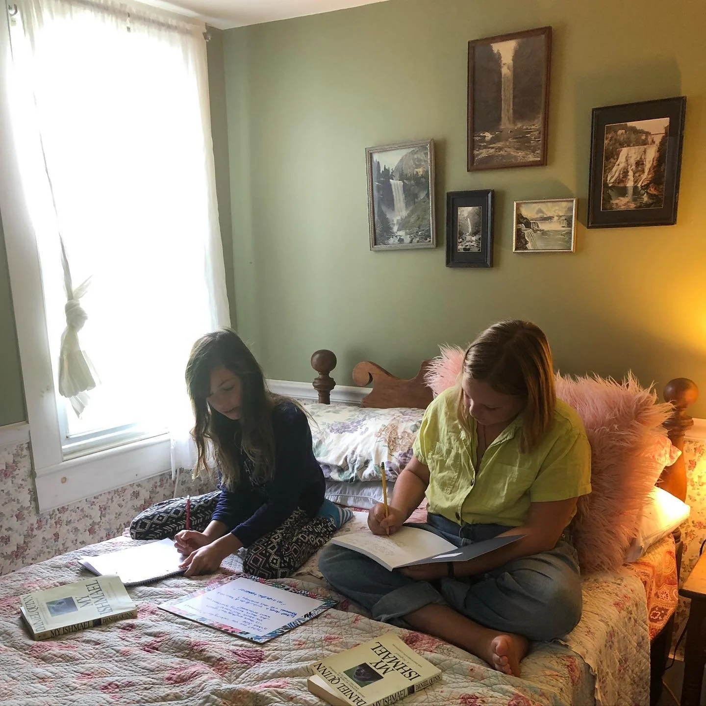 Two women sitting on a bed, reading and writing. The bed has floral bedding and pink fluffy pillows. There are books, notebooks, and whiteboards on the bed. The room has green walls, framed pictures of waterfalls, and a large window with white curtai