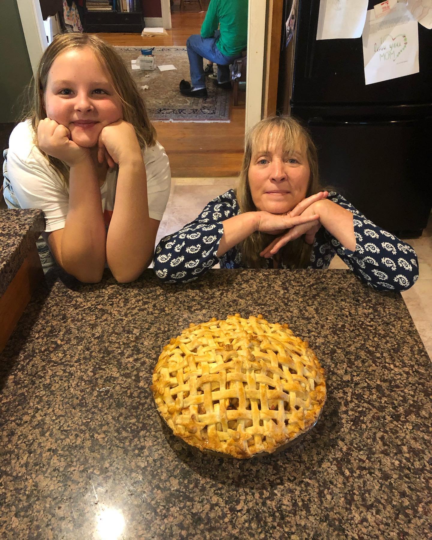 A young girl and an older woman are leaning on a kitchen counter behind a freshly baked apple pie with a lattice crust, smiling at the camera.