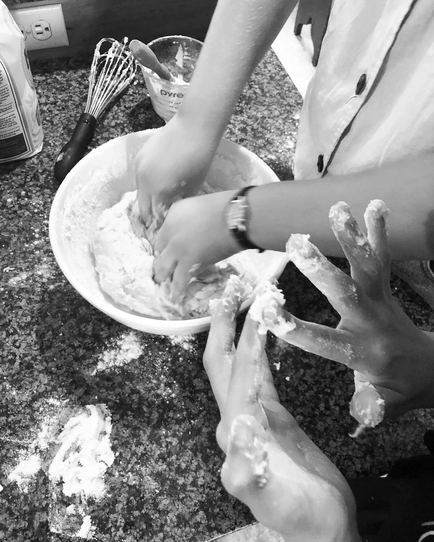 Two people are mixing dough in a large bowl on a kitchen counter. One person is using their hands, while the other is holding their hand. The countertop has flour scattered on it, and there is a whisk, a container with a wooden spoon, and a bottle of
