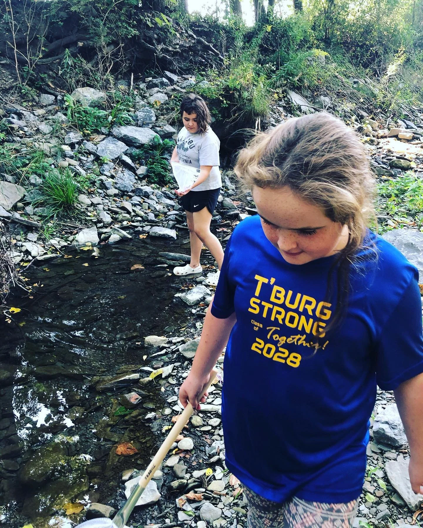 Two young girls explore a rocky creek in a wooded area, one holding a stick and the other looking into the water.