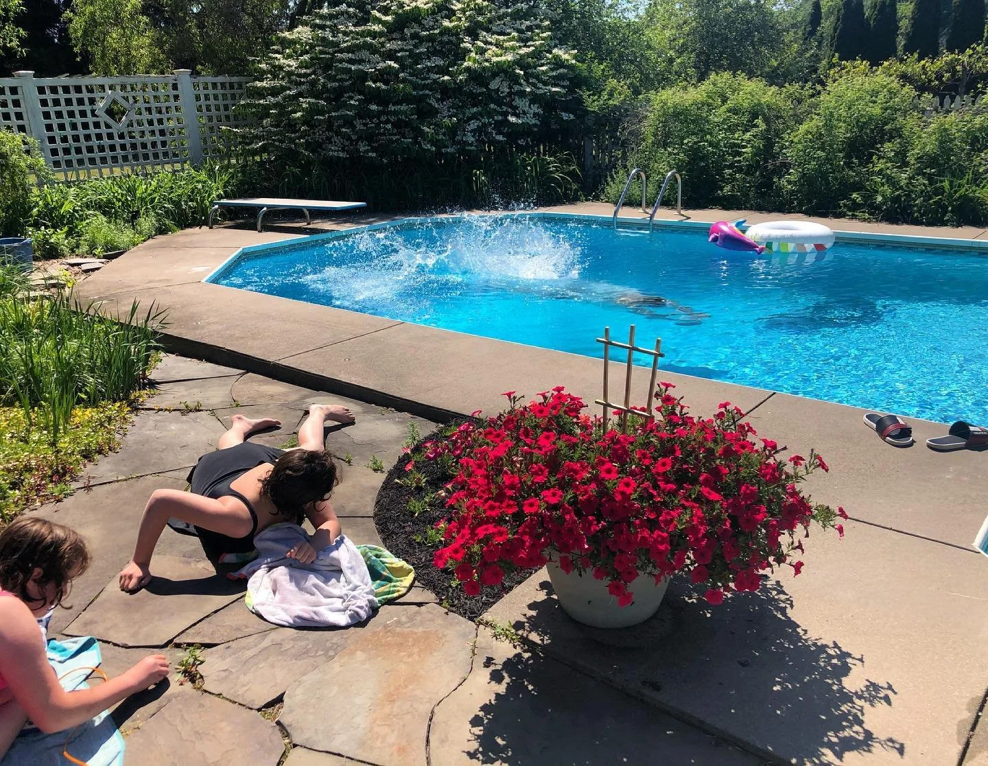 A backyard swimming pool with a splash occurring in the water. A ladder is visible at the far side of the pool, with a pink inflatable and a white float nearby. Two children are lying on the concrete patio near the pool, one with towels and the other