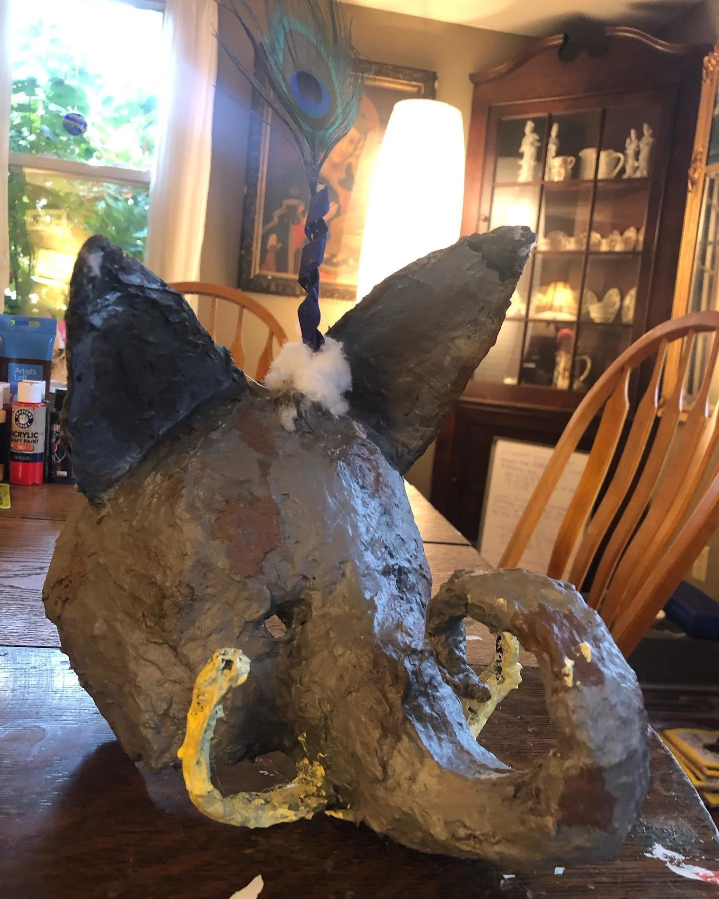 Handmade elephant sculpture with a peacock feather attached to its trunk, placed on a wooden table in a cozy room with wooden chairs, a window, and display cabinet in the background.