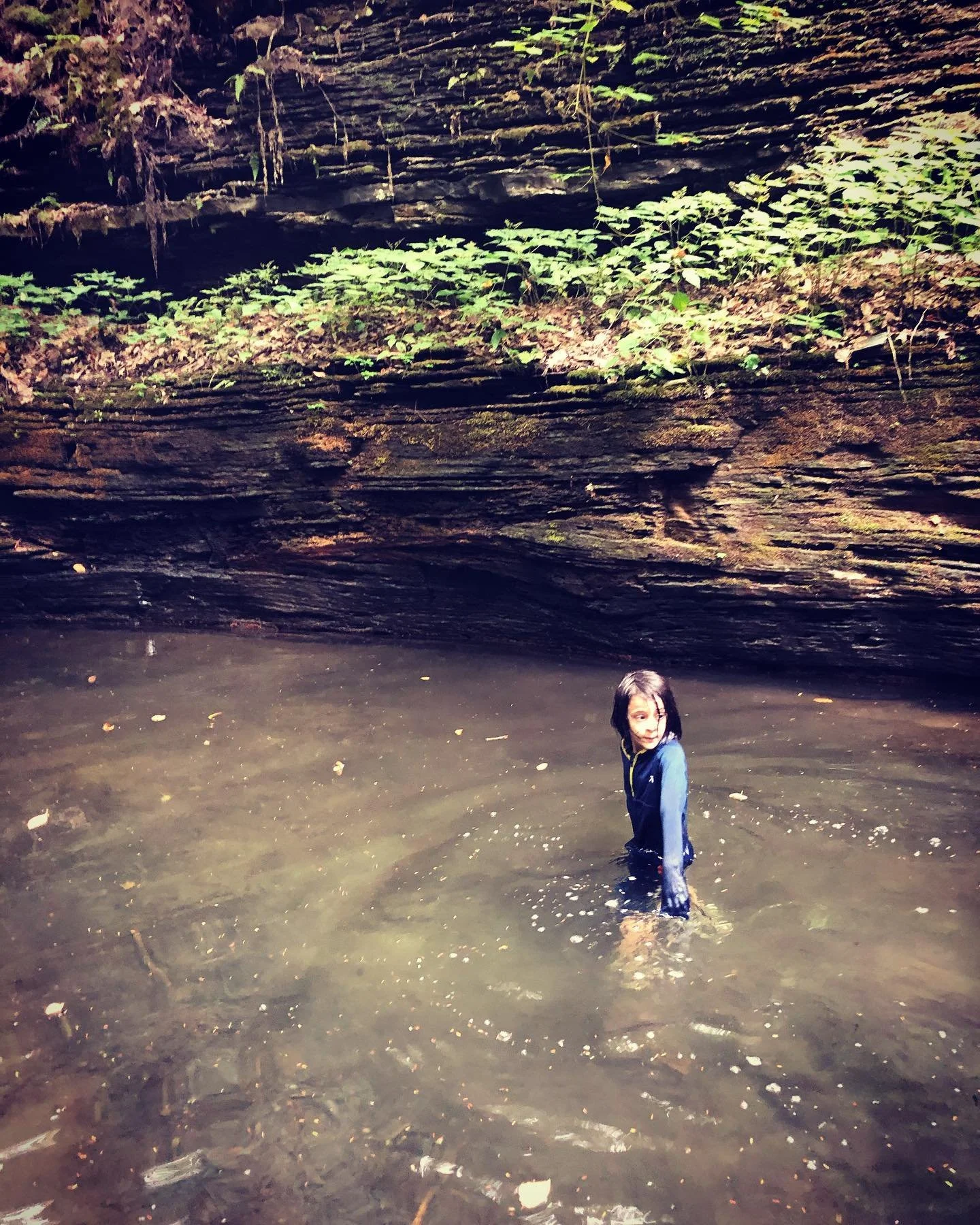 A young girl with dark hair, wearing a blue jacket, standing in a shallow muddy water pool at the base of a rock formation with green foliage on top.