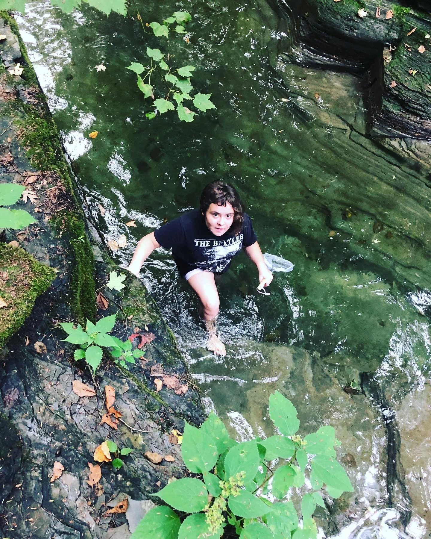 A woman standing in a creek surrounded by rocks and green foliage, looking up at the camera.