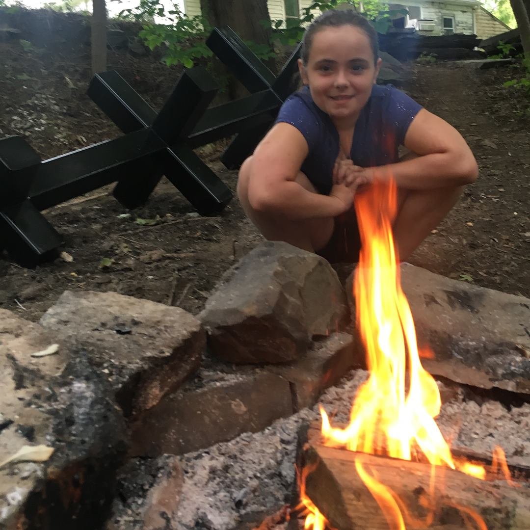 A girl crouching beside a campfire on the ground surrounded by rocks, with a black folding chair in the background in an outdoor setting with trees.