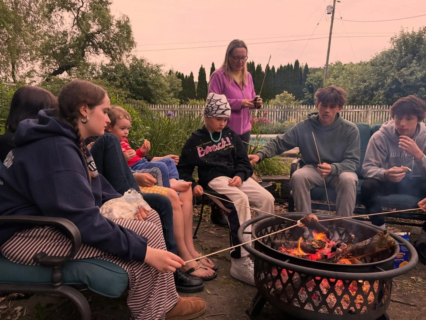 A group of children and teenagers sitting around a backyard firepit roasting marshmallows during sunset.