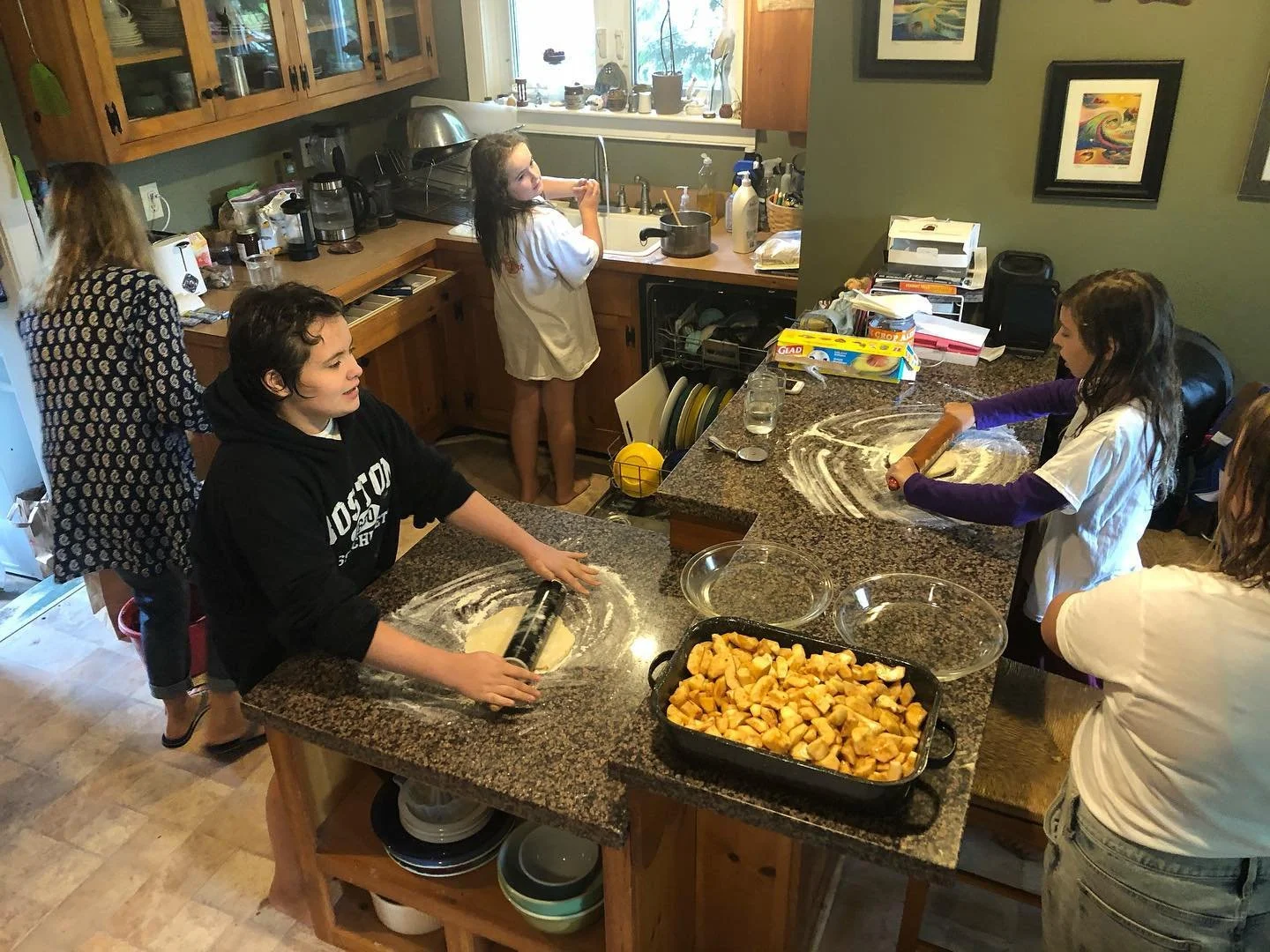 A group of children and a woman are baking cookies in a kitchen. They are rolling out dough with a rolling pin and have a tray of cubed cookie dough on the counter. The kitchen has wooden cabinets and various appliances on the counters.
