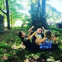 Two children and an adult sitting on a swing in a wooded area, enjoying a sunny day.
