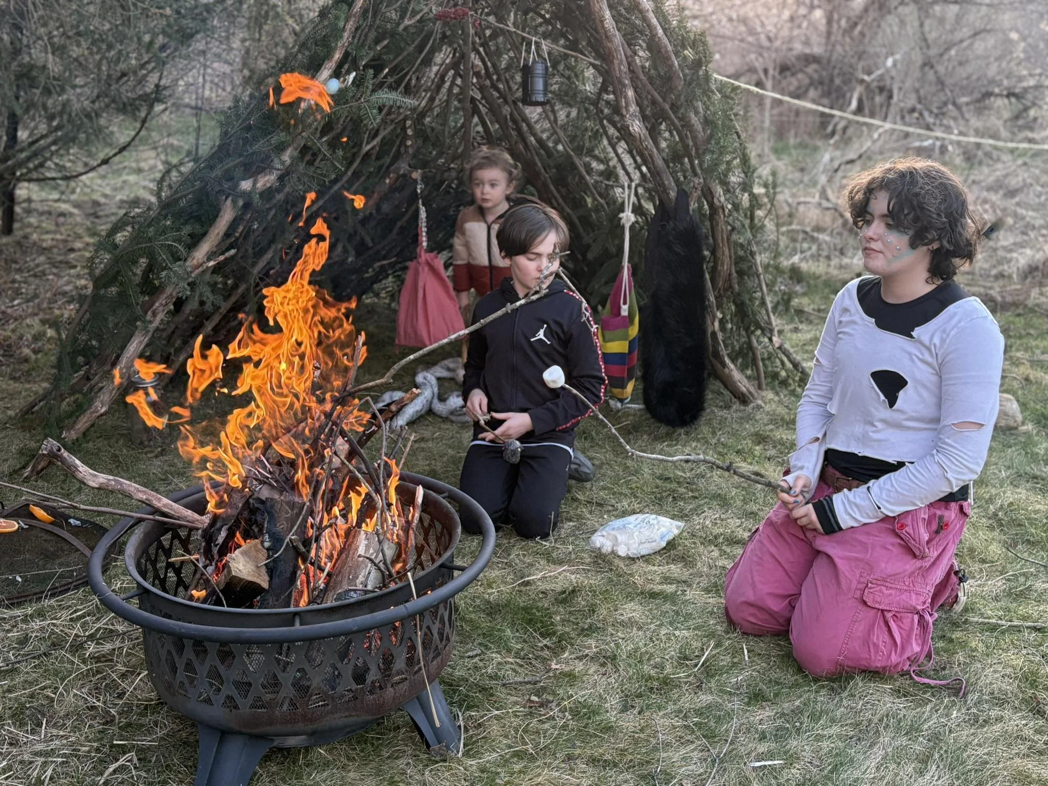Three children around a campfire inside a makeshift shelter made of branches and greenery, with some backpacks and clothing hanging nearby, in an outdoor setting.
