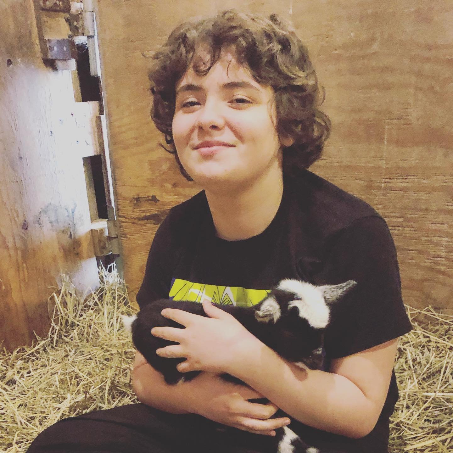 A smiling young person with curly brown hair holding a small black and white goat kid in their arms, sitting on straw in a wooden enclosure.