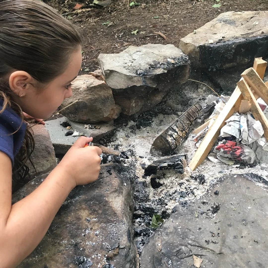 A girl crouching near a campfire with ashes, burnt wood, and crumpled paper surrounded by large rocks.