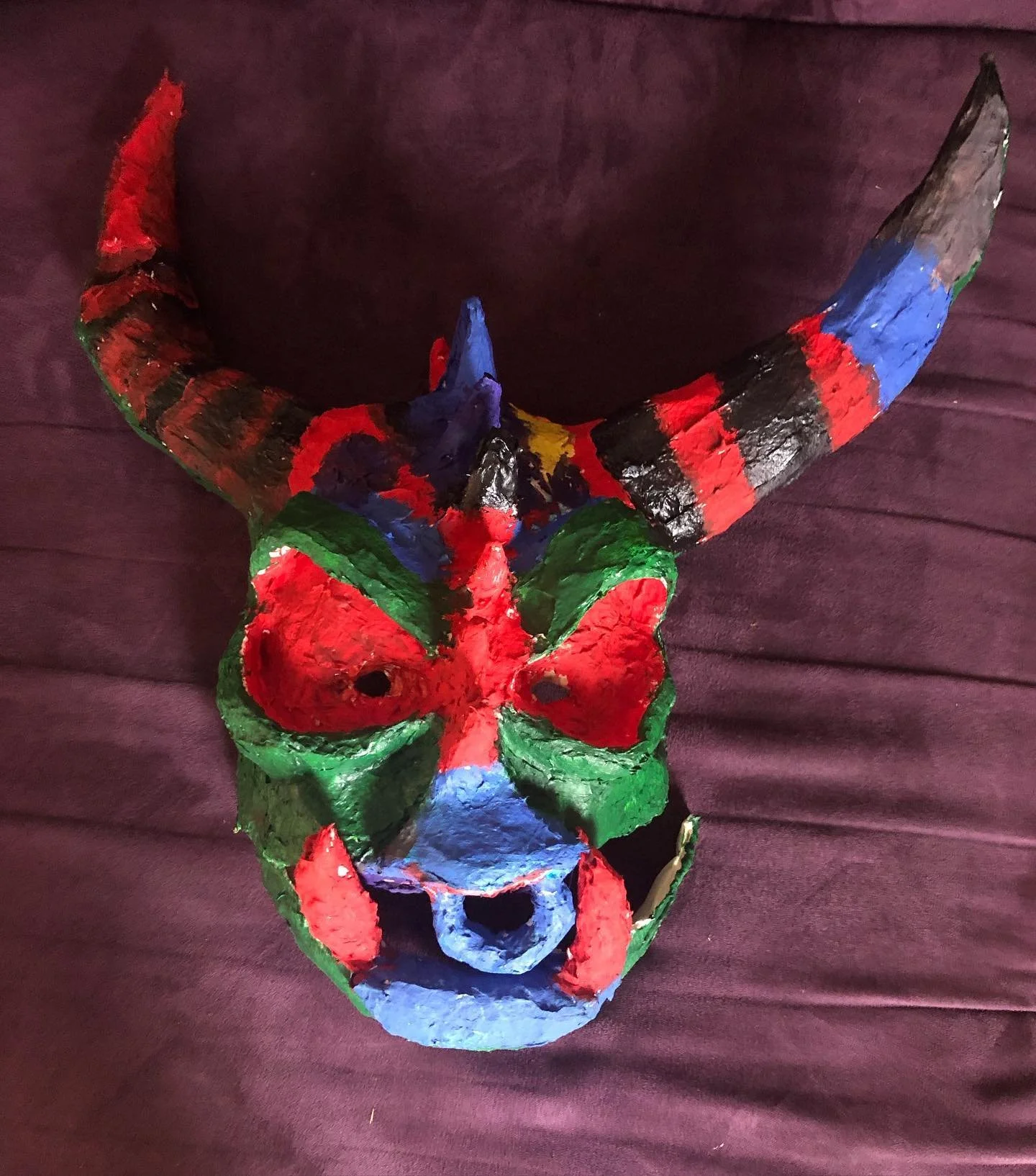 Colorful hand-crafted dragon mask with large horns, painted in red, green, blue, yellow, and black, placed on a dark purple fabric background.