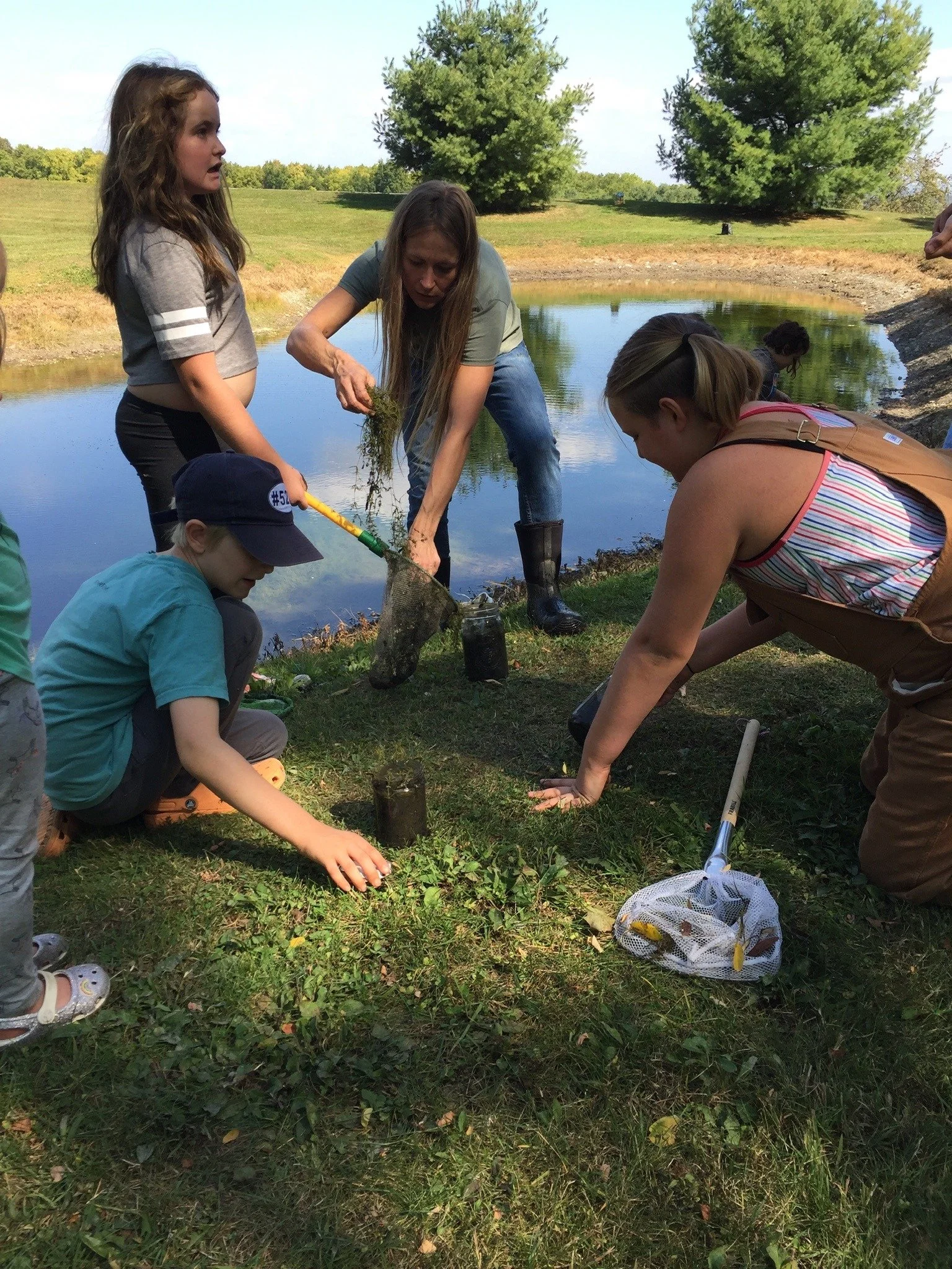 A group of people, including children and adults, planting trees by a pond in an outdoor setting.