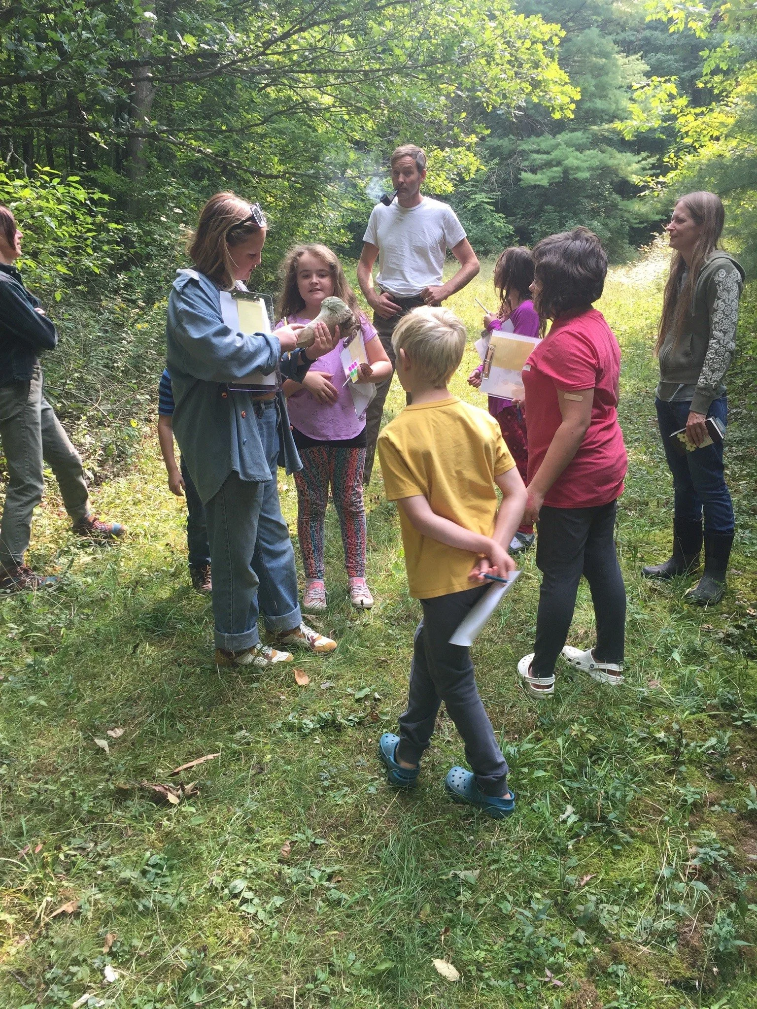 Group of children and adults in a forest clearing, participating in an outdoor educational activity or nature walk, with some children holding clipboards and notebooks.