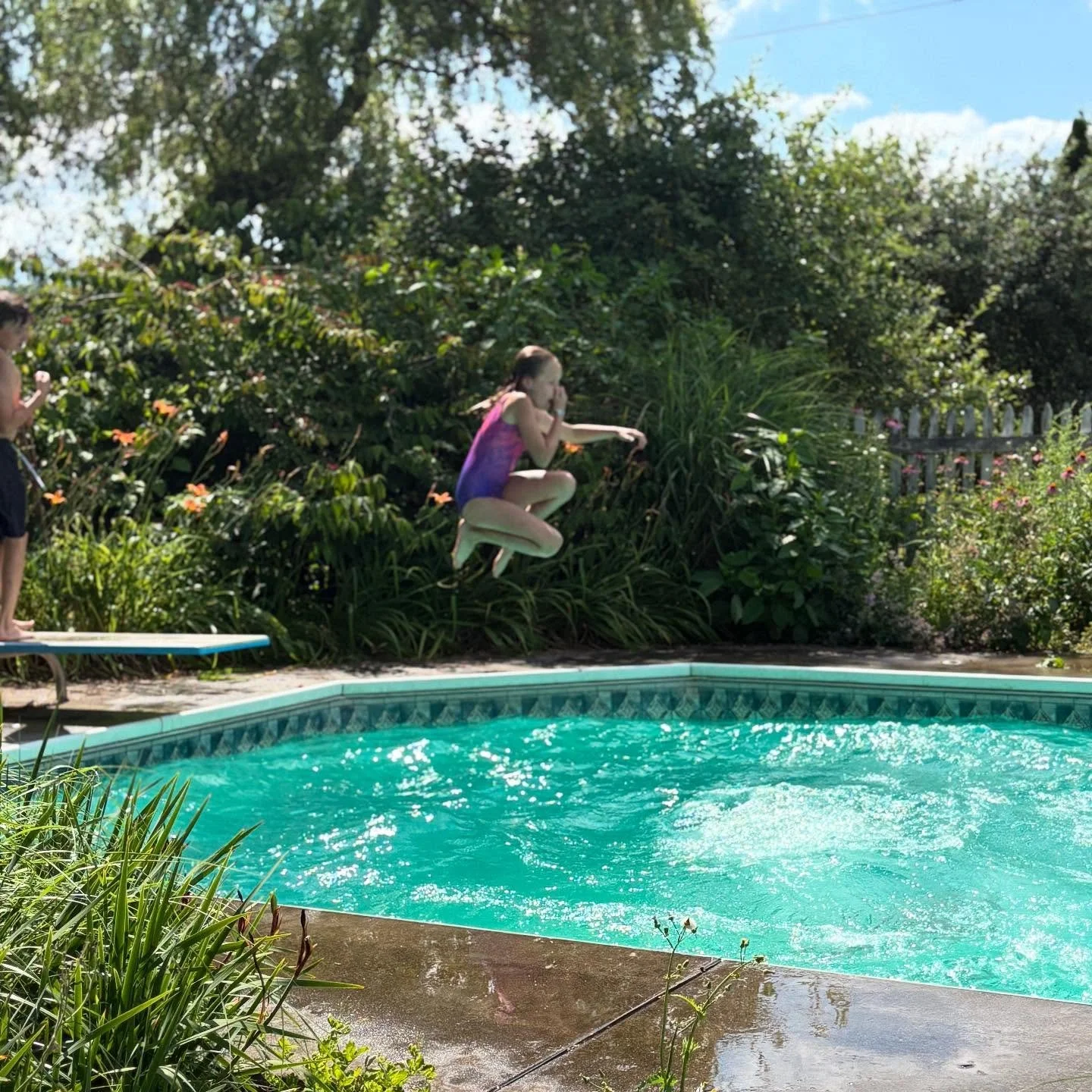 A young girl jumping into a backyard swimming pool as another child stands nearby on the pool deck. The area is surrounded by lush green plants and a white picket fence, with blue sky visible in the background.