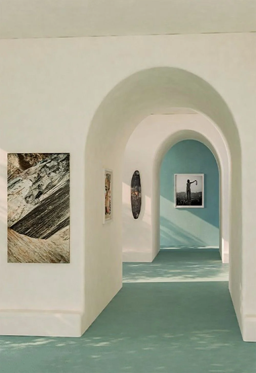 Interior of an art gallery with arched doorways, displaying abstract paintings and photographs on white walls, with sunlight casting shadows on the light blue and teal-colored walls and greenish floor.