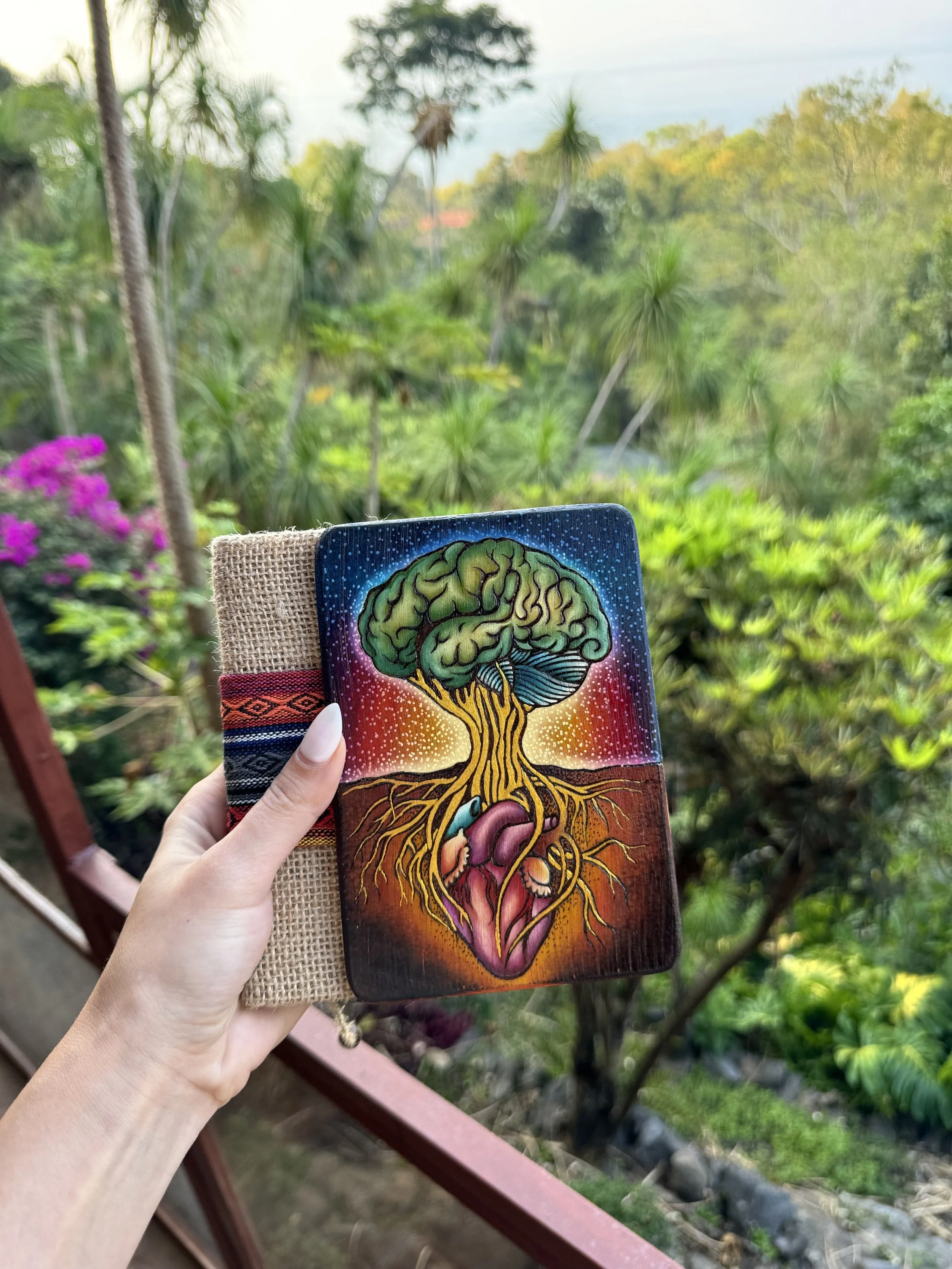 A hand holding a tarot card with a colorful illustration of a brain, trees, and a heart, against a lush green outdoor background.
