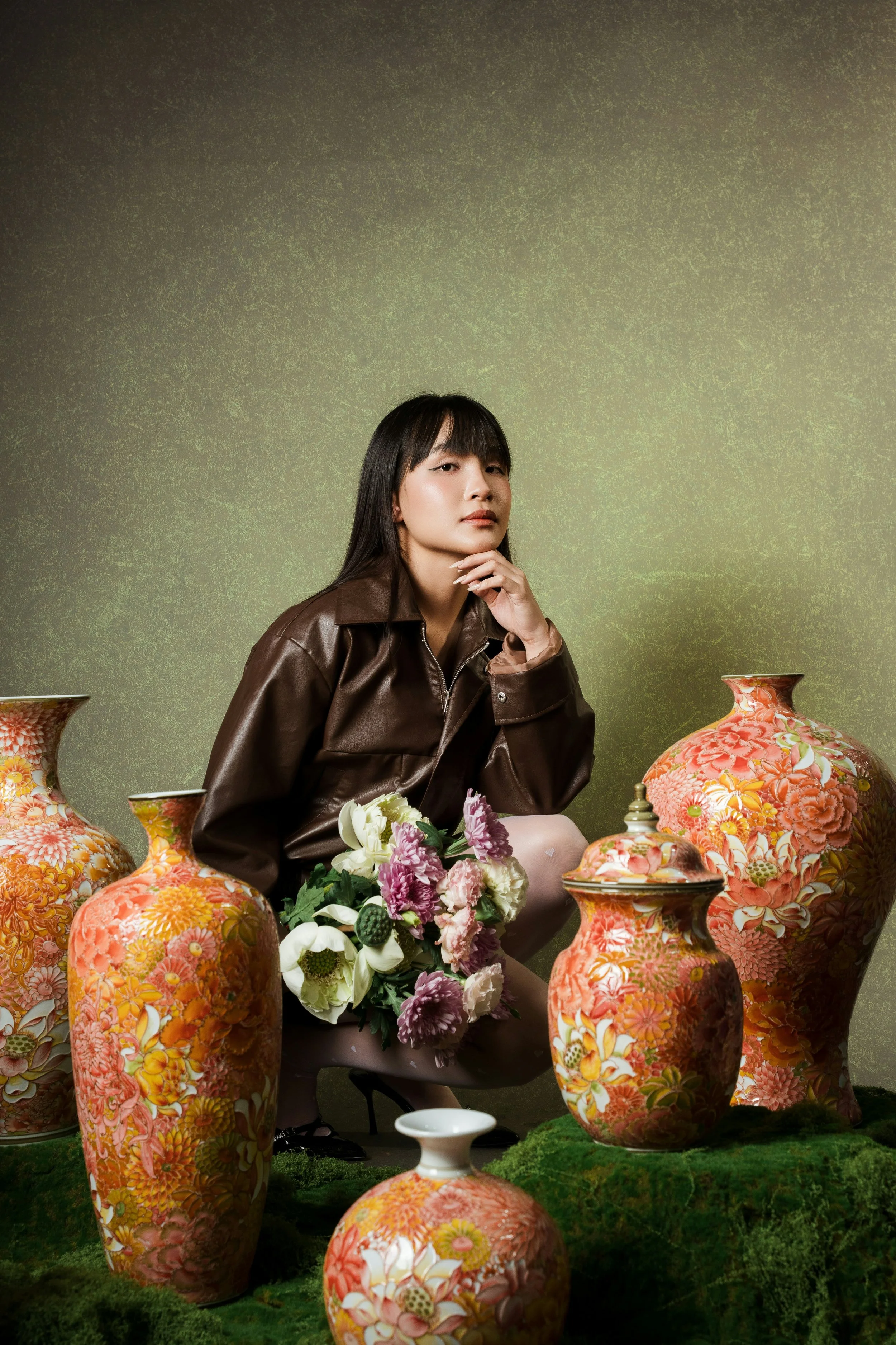A woman sitting among large, colorful floral vases, holding a bouquet of flowers with flowers on the ground and a green textured backdrop.