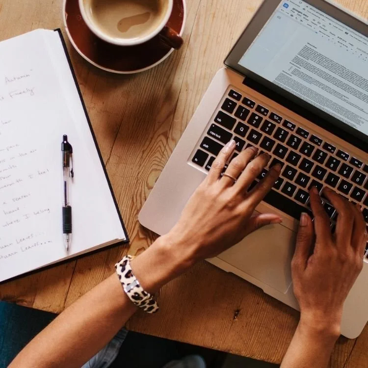 A person working on a laptop at a wooden table with a notebook, pen, and a cup of coffee.