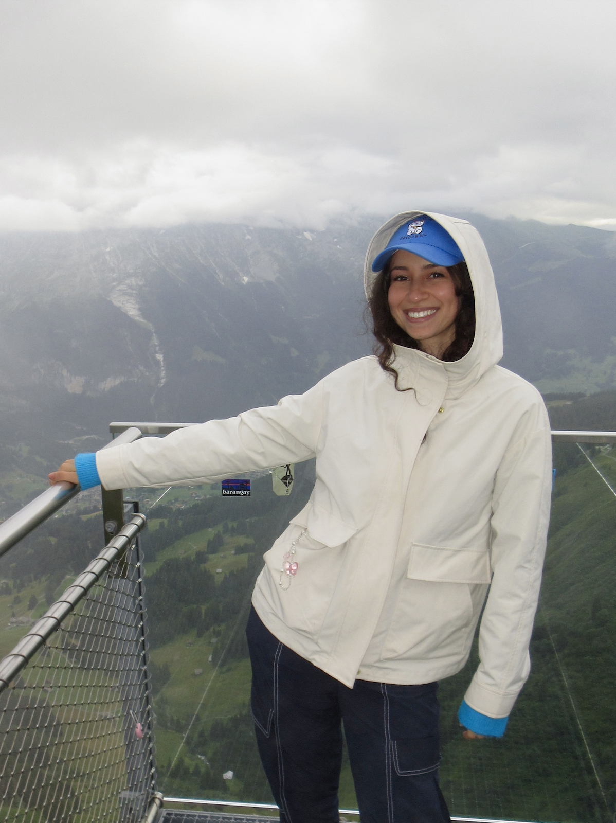 A woman wearing a white jacket, a blue cap, and dark pants standing on a viewing platform outdoors with mountains and cloudy sky in the background, smiling at the camera.