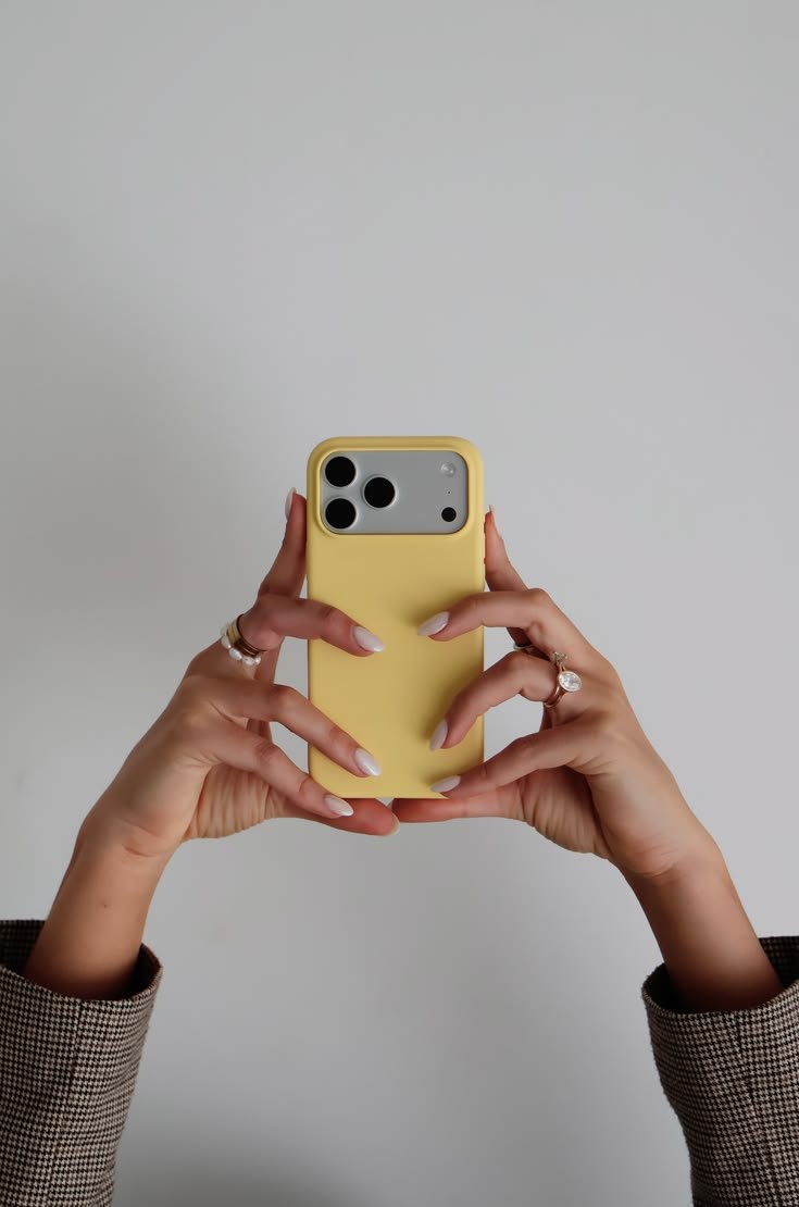 Person taking a selfie with a yellow phone case, holding the phone with both hands. The person is wearing rings and a brown checkered blazer, against a plain gray background.