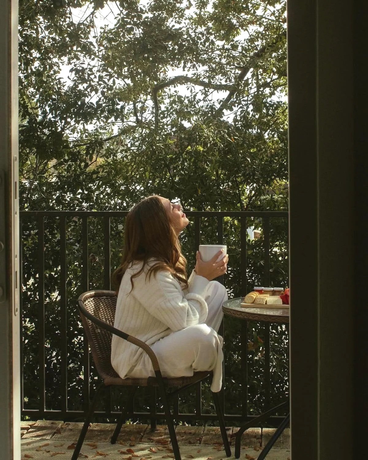 A woman with long brown hair, wearing a white sweater, sitting on a balcony, holding a white mug, with a table nearby that has a plate of cookies and jam jars, surrounded by greenery and trees.