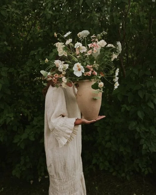 Person in a beige dress holding a large vase with a bouquet of assorted flowers, standing outdoors against a background of green foliage.