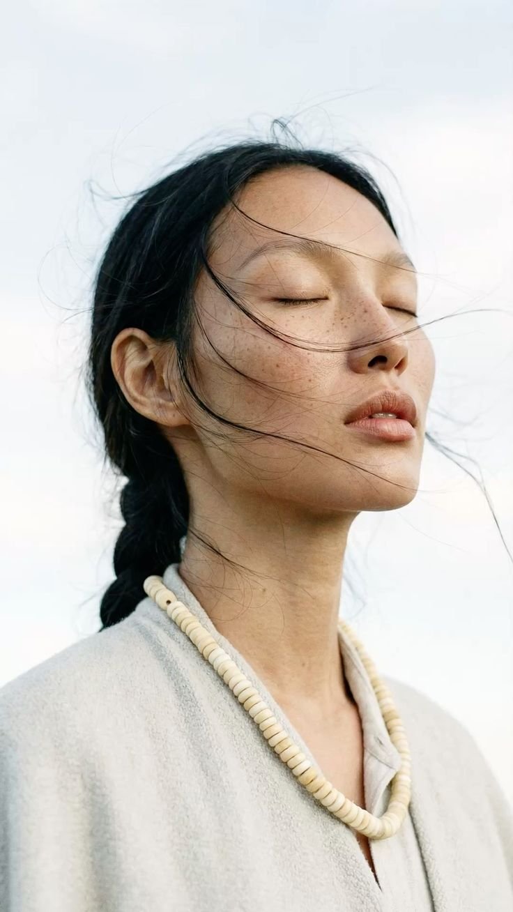 A woman with long black hair, lightly freckled face with closed eyes, wearing a white beaded necklace and a light-colored top, outdoors with a cloudy sky background.