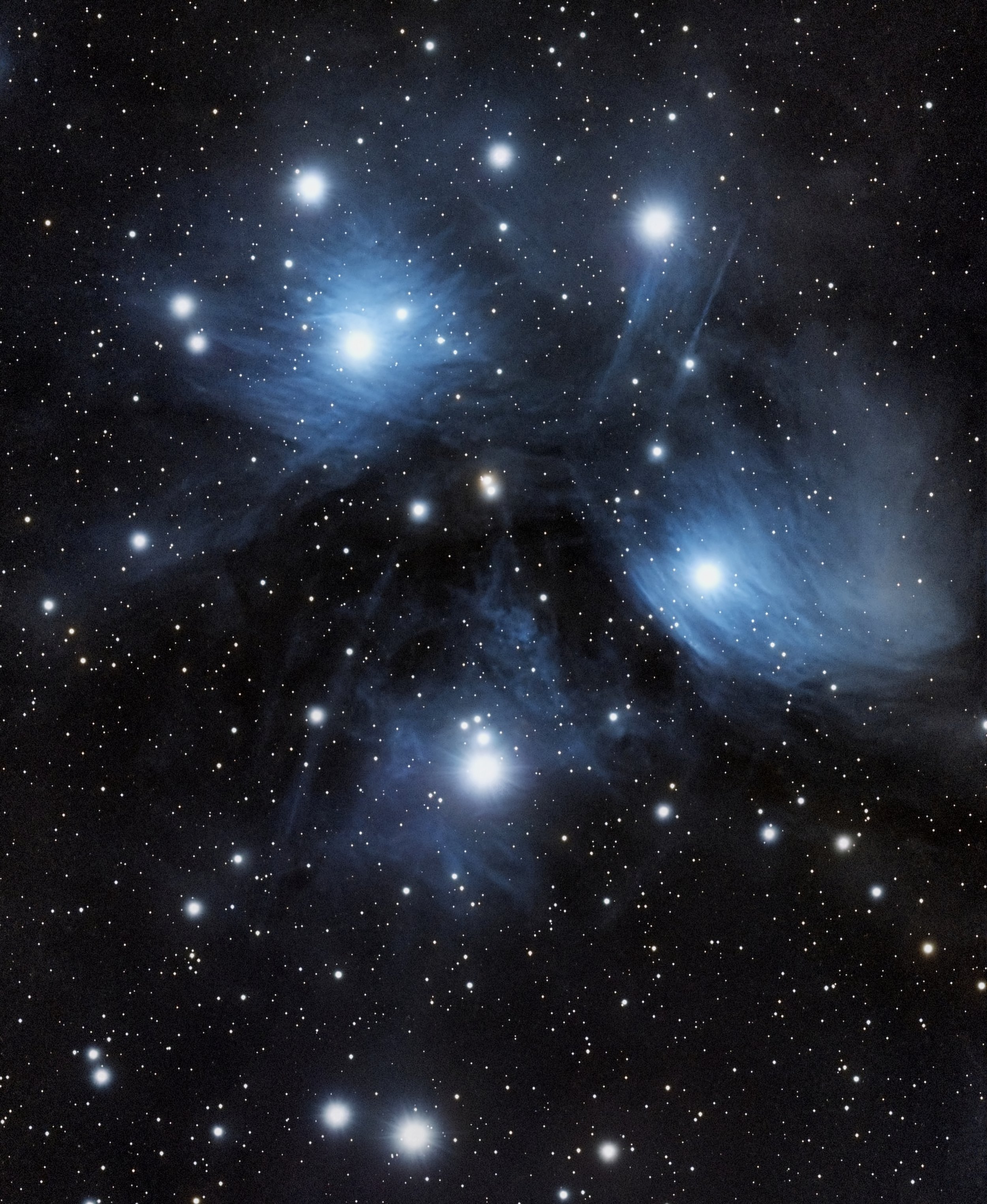A detailed photograph of The Pleiades asterism, showing numerous stars, and blue reflection nebulosity with a dark background.