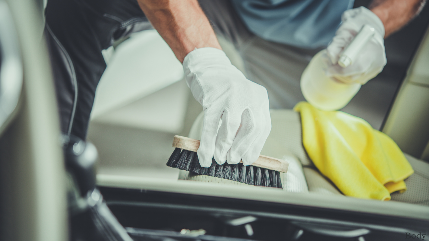 Person in gloves cleaning a car seat with a brush, yellow cloth, and spray bottle.