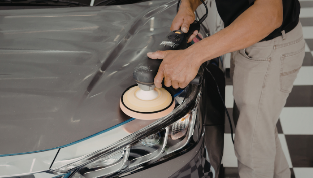 Person polishing the hood of a black car with a handheld electric polisher in a garage.
