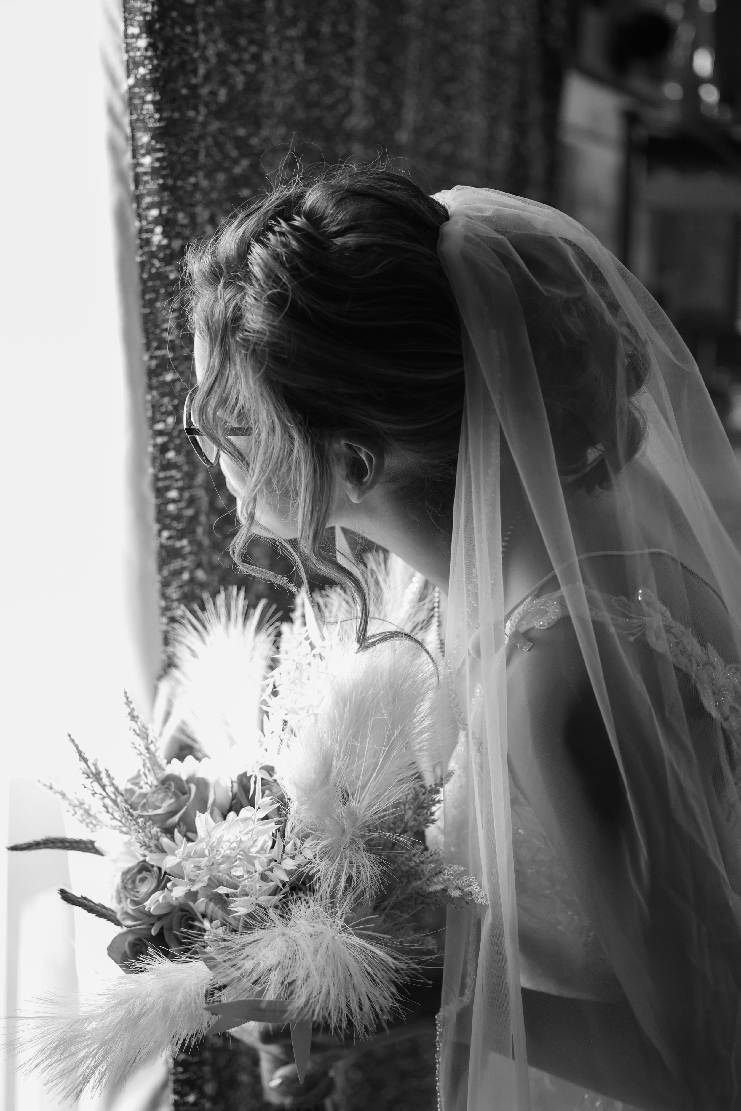 Black and white photo of a bride holding a bouquet of flowers, wearing a veil, with a blurred outdoor background.