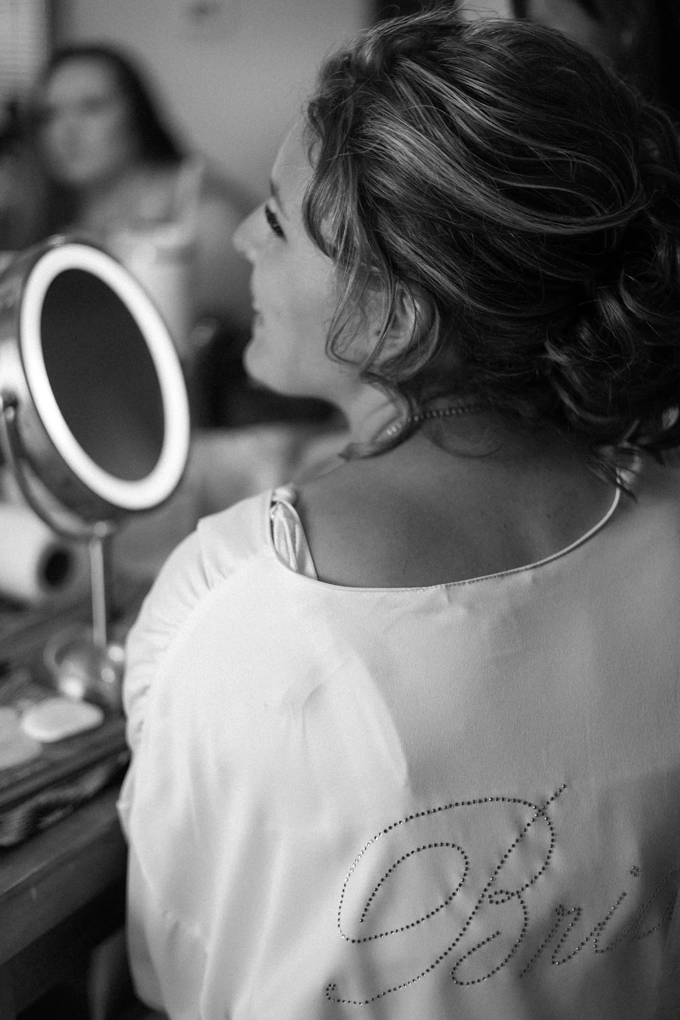 A woman with curly hair applies makeup in front of a mirror. She is wearing a light-colored shirt with a decorative design or text on it. In the background, another person is slightly out of focus.