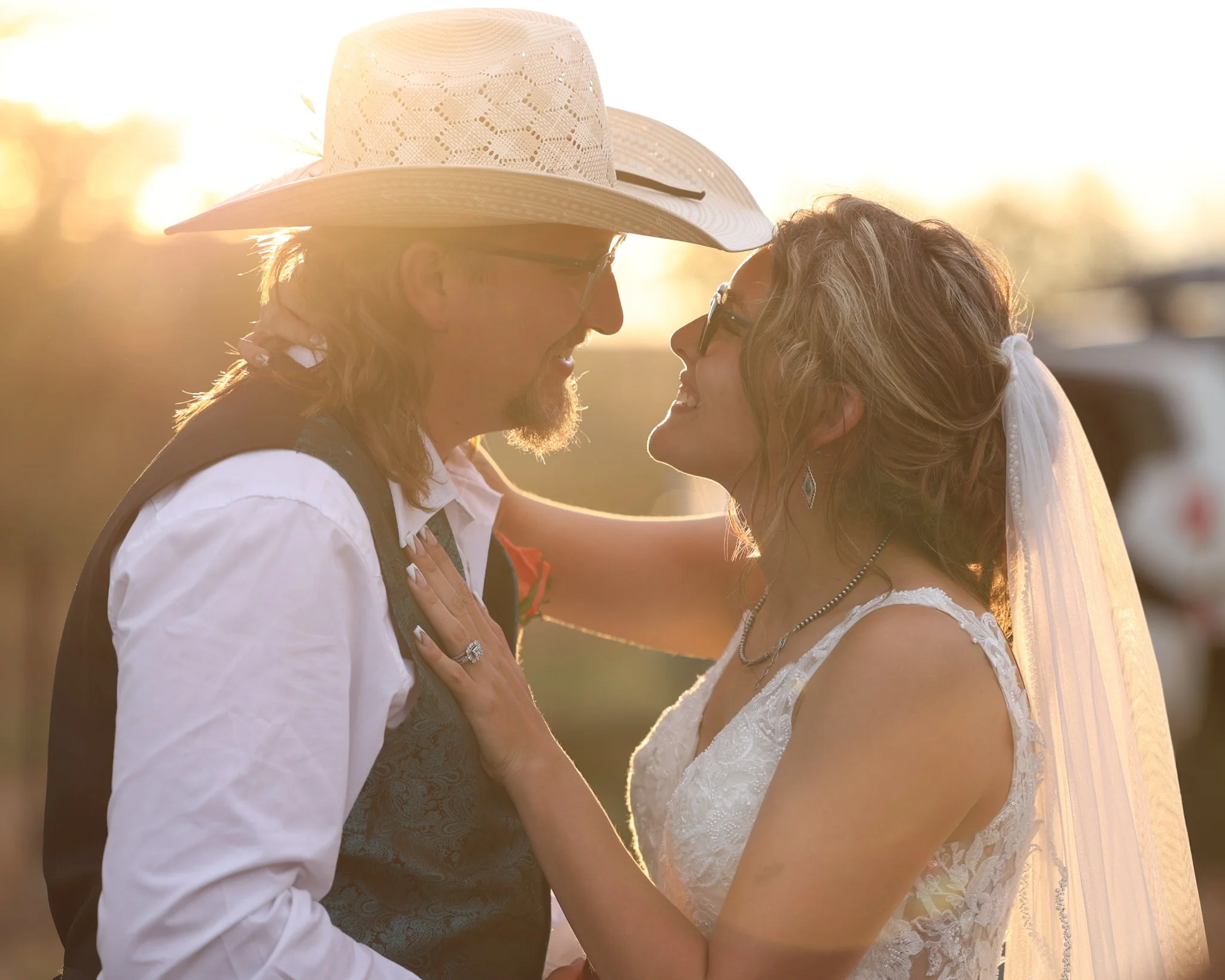 A couple is about to kiss outdoors during sunset, with the man wearing a wide-brimmed hat and glasses, and the woman wearing glasses and earrings.