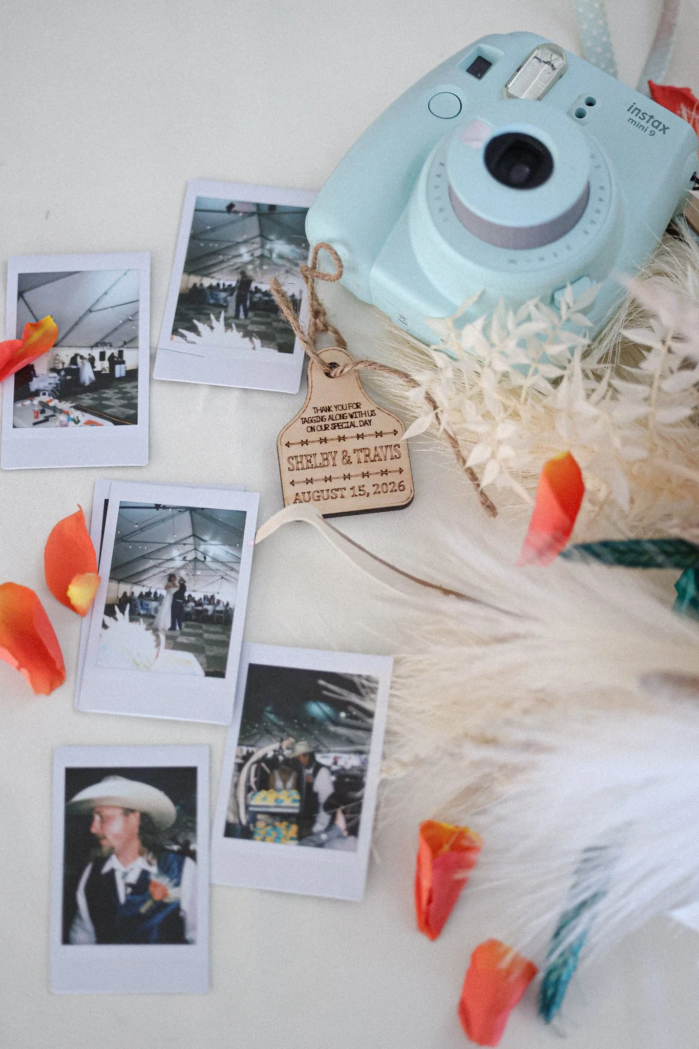 Polaroid photographs scattered on a surface, with a small sign that reads Shelby & Travis, August 15, 2026, attached with a string. A pastel blue Instax Mini 9 camera sits amidst decorative paper and paper flowers.