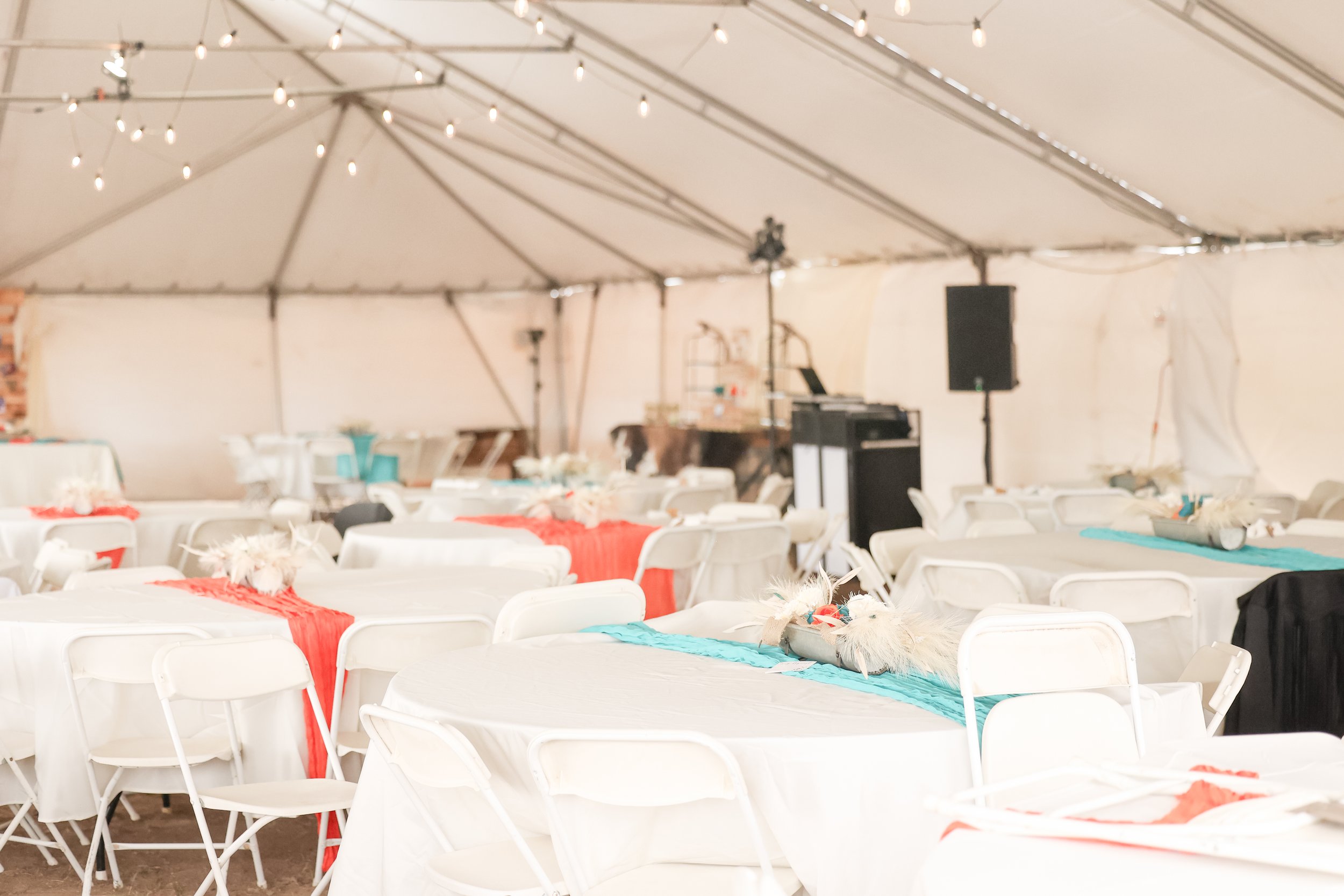 Inside of a large event tent decorated with string lights, with round tables and white chairs. The tables are decorated with red and blue table runners and centerpieces with feathers.